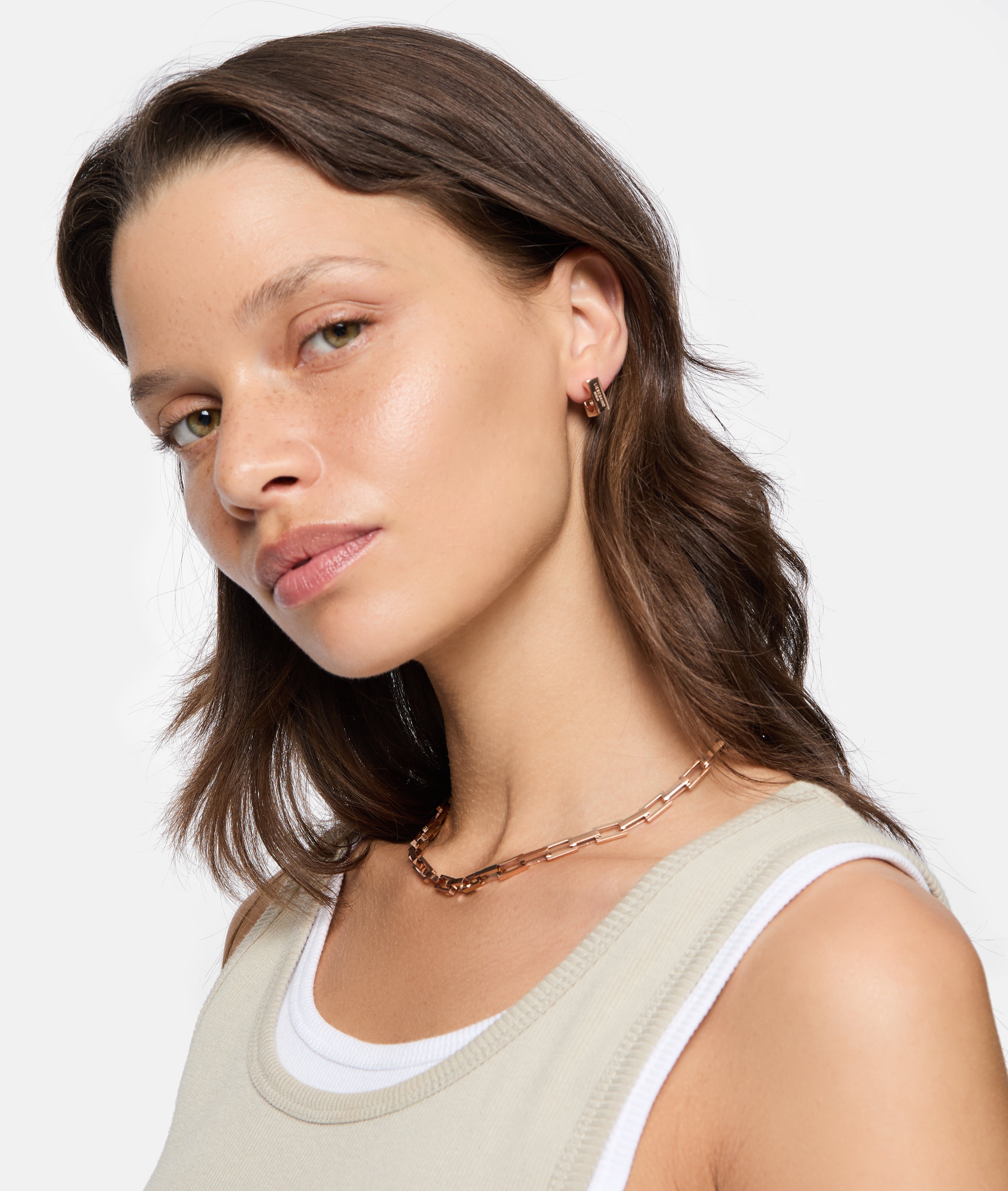 Frau mit gewellten braunen Haaren trägt ein beiges Tanktop, goldene Ohrringe und die LIEBESKIND BERLIN Halskette - Urban Chic von Liebeskind Berlin.