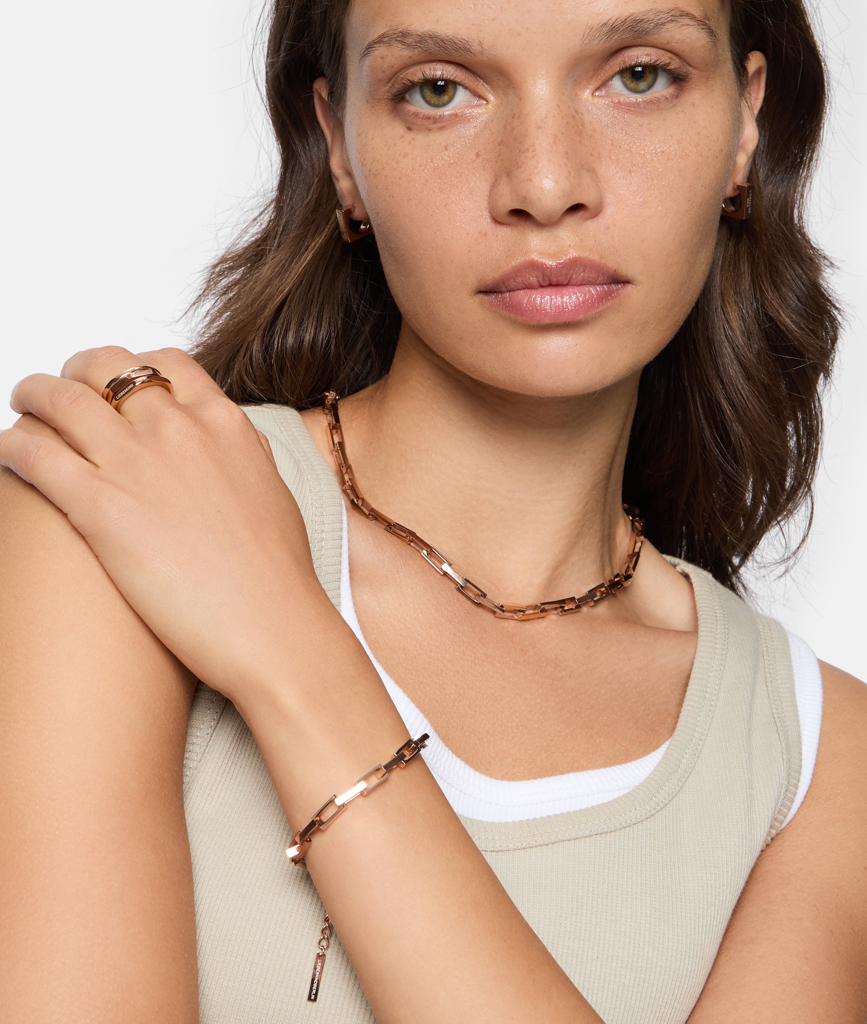 Eine Frau in einem beigen Tanktop über einem weißen Hemd modelliert Goldschmuck, darunter Ohrringe, eine Halskette, ein Armband und den Liebeskind Berlin Urban Chic Ring (Größe 58).