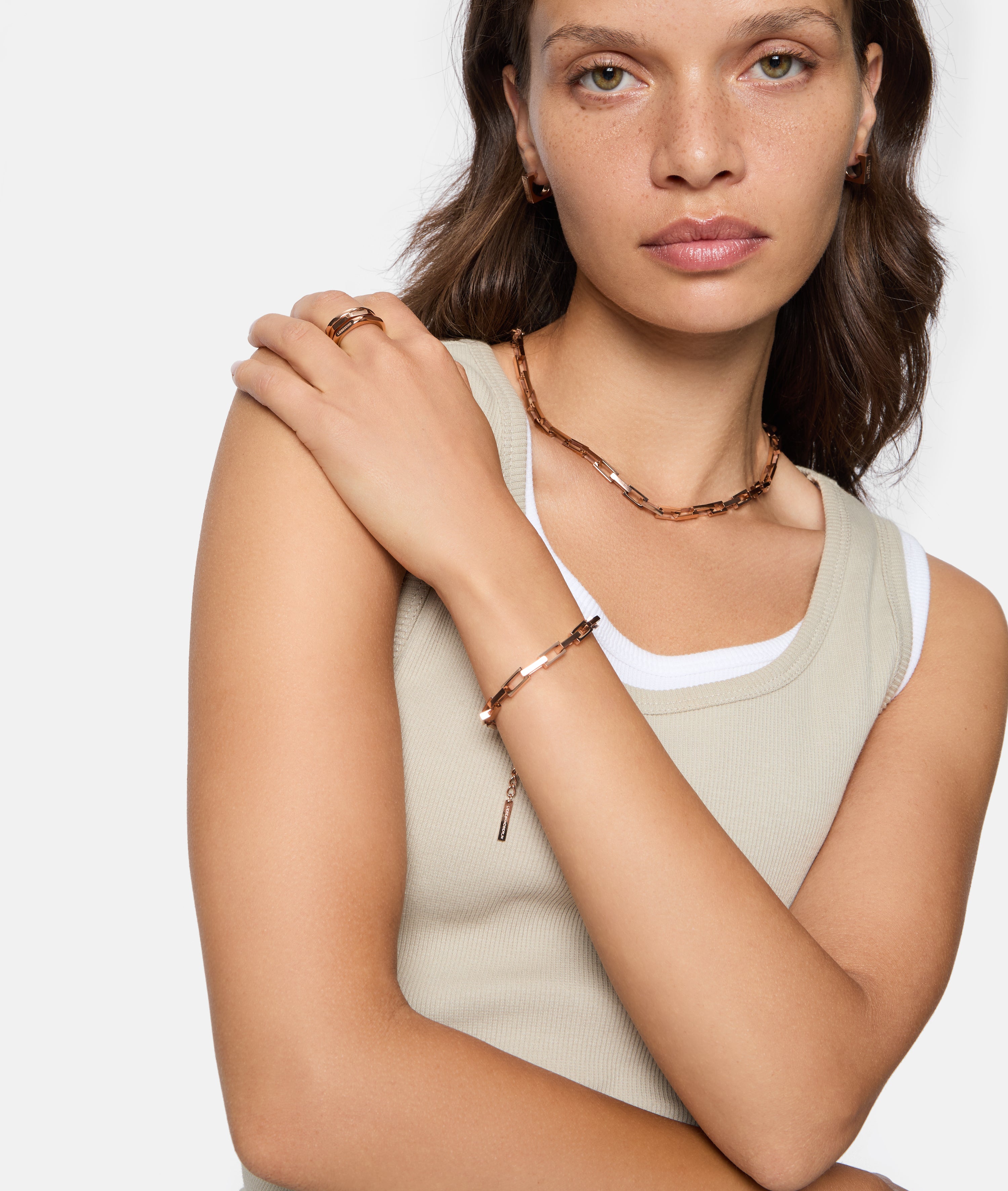 Eine Frau in einem beigen Tanktop trägt goldenen Schmuck, darunter das Liebeskind Berlin Urban Chic Armband, eine Halskette, einen Ring und kleine Ohrringe für einen urbanen Look.