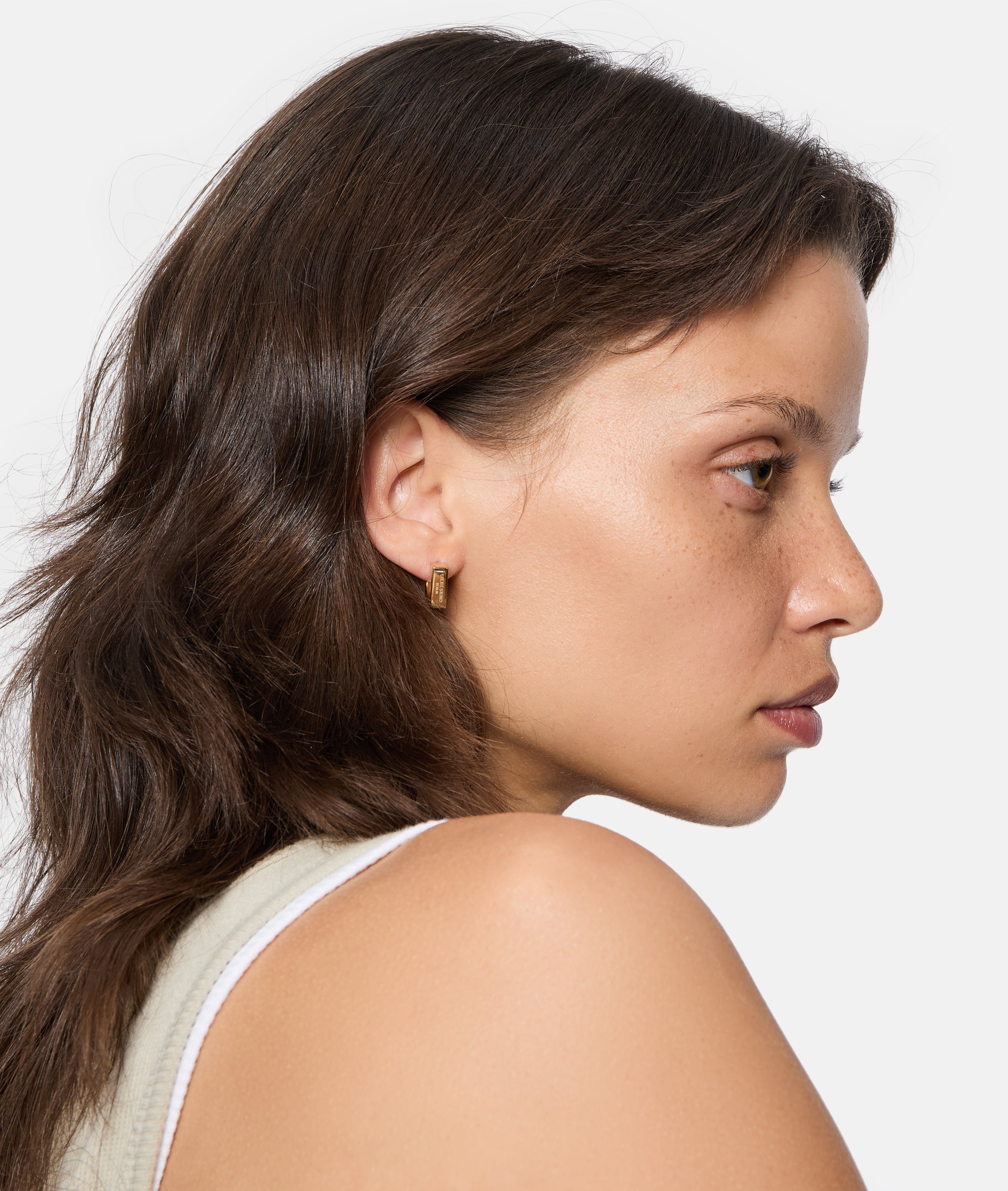 Frau mit braunen Haaren im Profil trägt ein ärmelloses Oberteil und stilvolle Liebeskind Berlin Creole - Urban Chic Ohrringe, ein Must-Have-Accessoire, vor einem weißen Hintergrund.