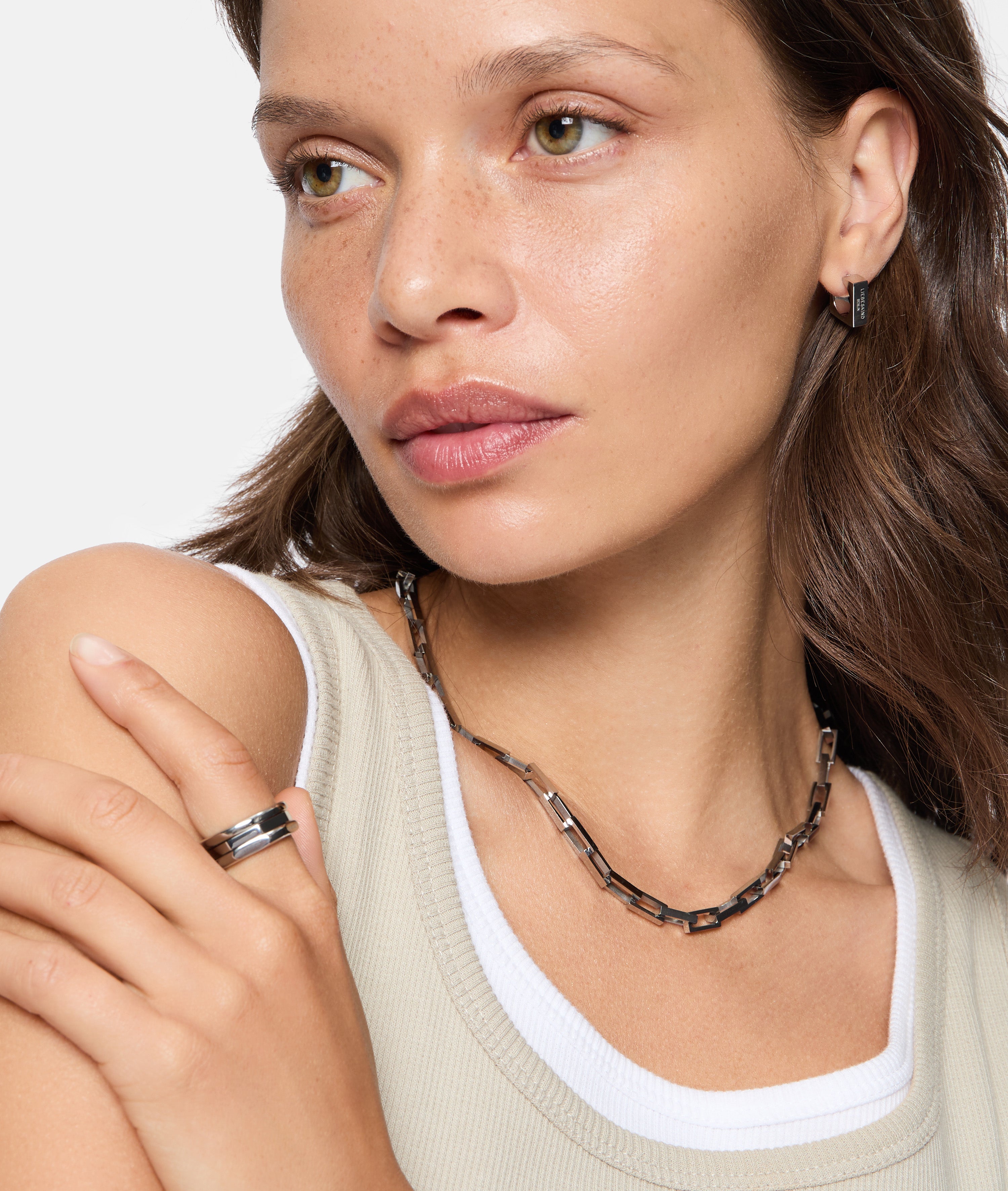 Frau trägt ein ärmelloses Oberteil, einen Liebeskind Berlin Urban Chic Ring in Größe 56, gepaart mit einer Kettenhalskette und Ohrringen, Blick zur Seite.