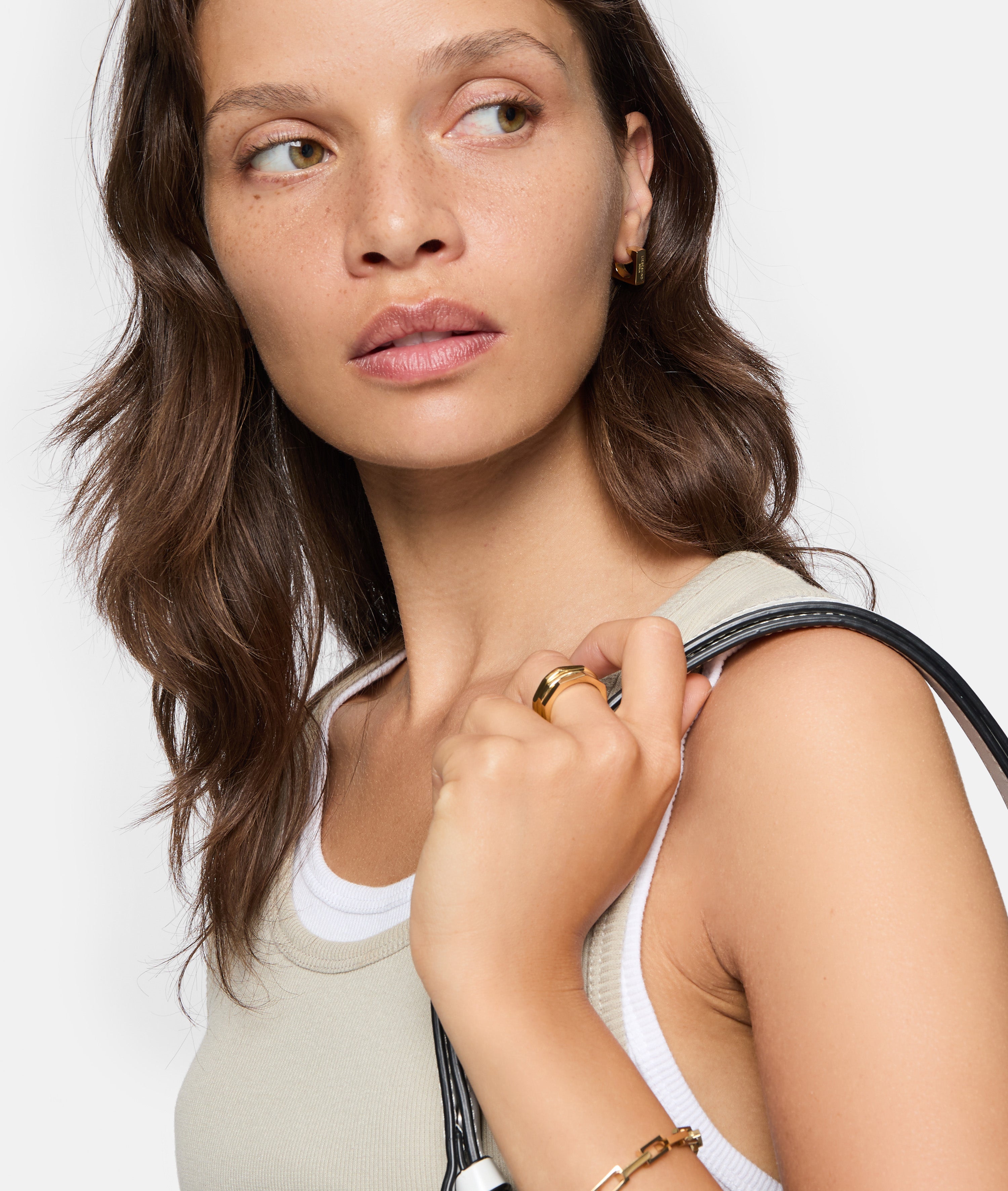 Eine Frau mit gewellten braunen Haaren in einem Tanktop und Goldschmuck trägt den LIEBESKIND BERLIN Ring - Urban Chic in 54 von Liebeskind Berlin und hält ein schwarzes Armband über ihre Schulter.