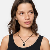 Frau mit gewellten braunen Haaren, bekleidet mit einem schwarzen Tanktop, trägt die LIEBESKIND BERLIN Halskette - The T-Bar Plate von Liebeskind Berlin mit einem schwarzen, achteckigen Anhänger, schaut zur Seite.