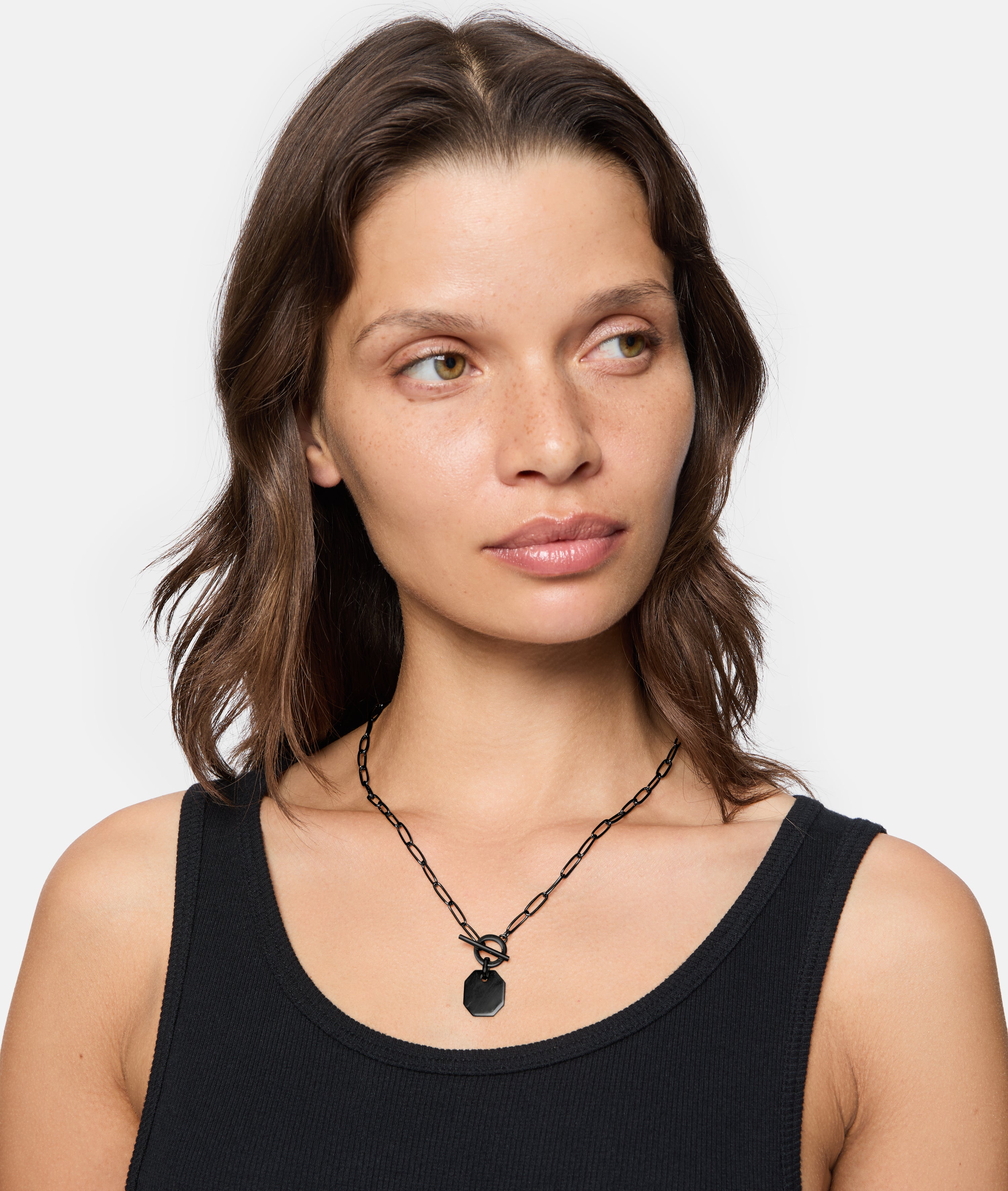 Frau mit gewellten braunen Haaren, bekleidet mit einem schwarzen Tanktop, trägt die LIEBESKIND BERLIN Halskette - The T-Bar Plate von Liebeskind Berlin mit einem schwarzen, achteckigen Anhänger, schaut zur Seite.