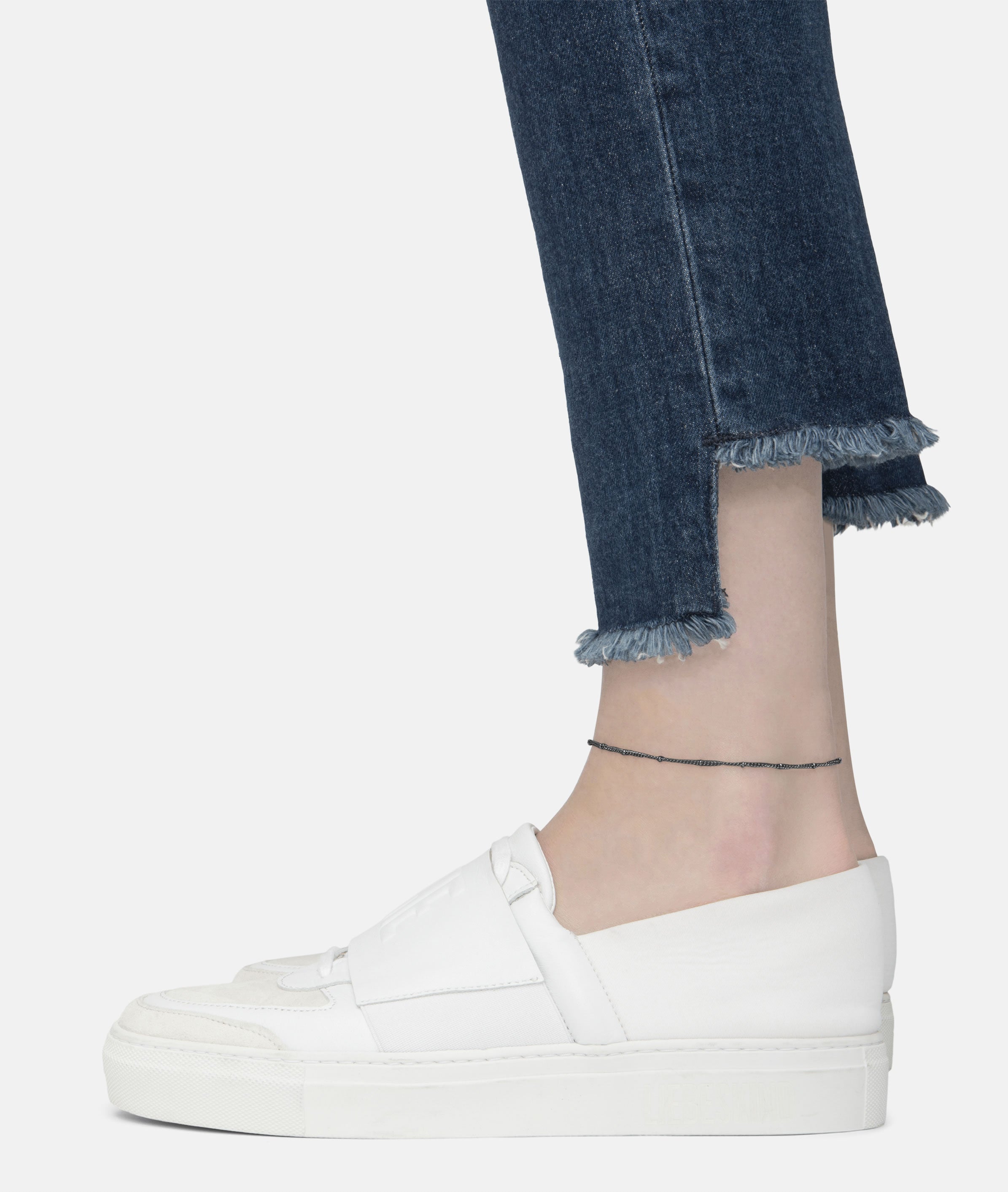 Eine Person trägt eine Jeans mit ausgefranstem Saum, weiße Slipper und die LIEBESKIND BERLIN Fußkette - The Simple Solo von Liebeskind Berlin vor einem schlichten Hintergrund.