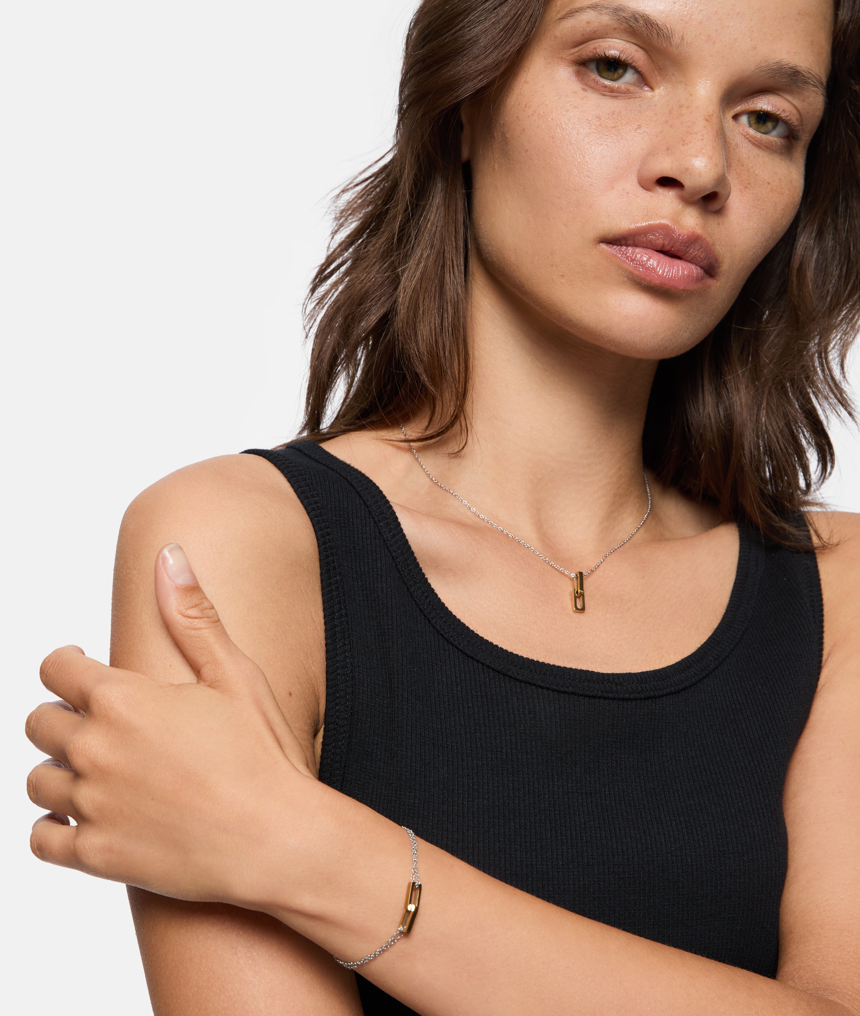 Frau in schwarzem Tank-Top trägt ein Liebeskind Berlin Armband - Mini Link und eine Goldbarren-Halskette mit rechteckigem Anhänger, schaut in die Kamera.