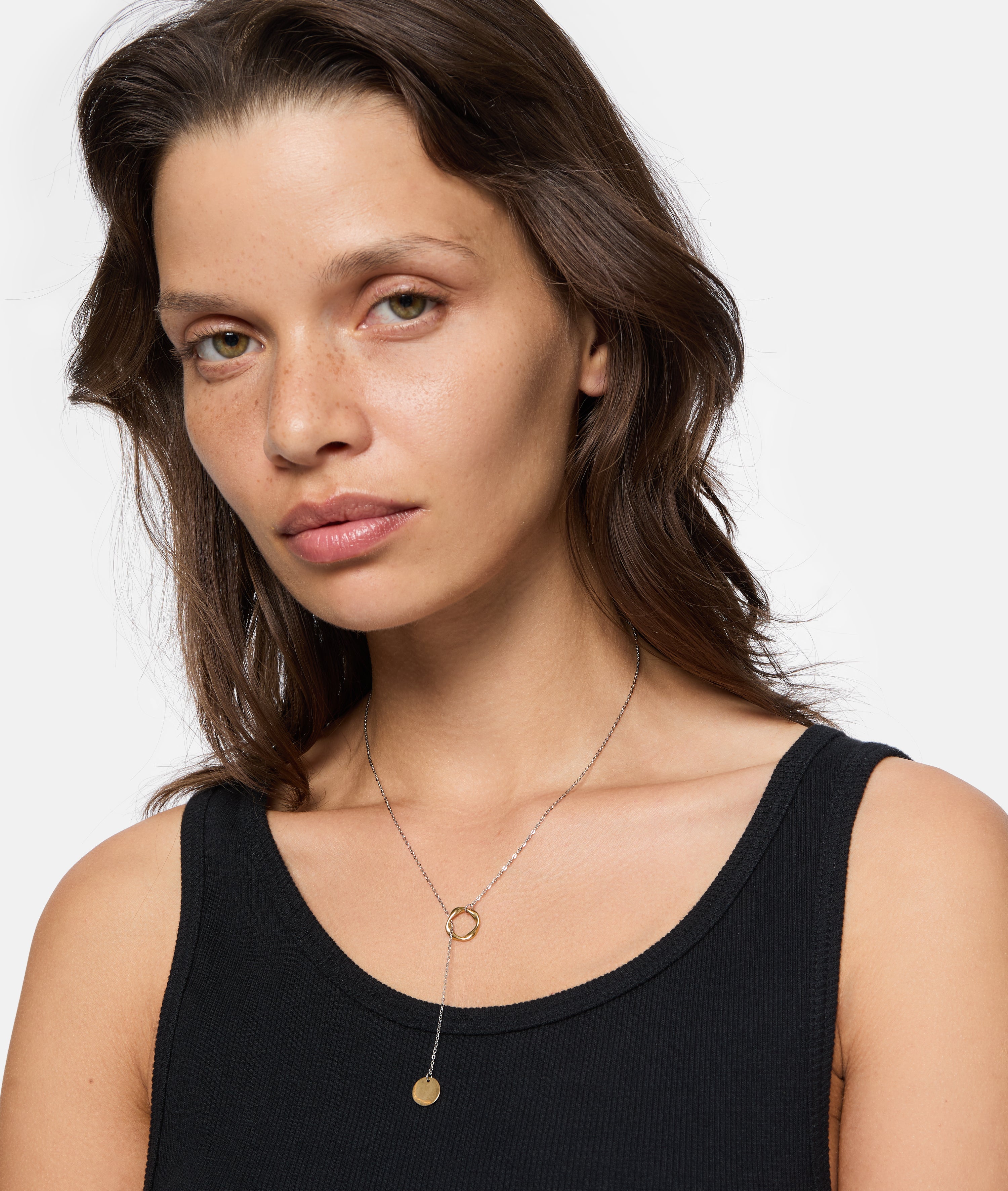 Frau mit gewelltem braunem Haar trägt ein schwarzes Tank-Top und die LIEBESKIND BERLIN Halskette - The Organic Twist, posiert vor einem weißen Hintergrund. Ein stilvolles Beispiel für die Schichtung von Gold- und Edelstahl-Halsketten von Liebeskind Berlin.