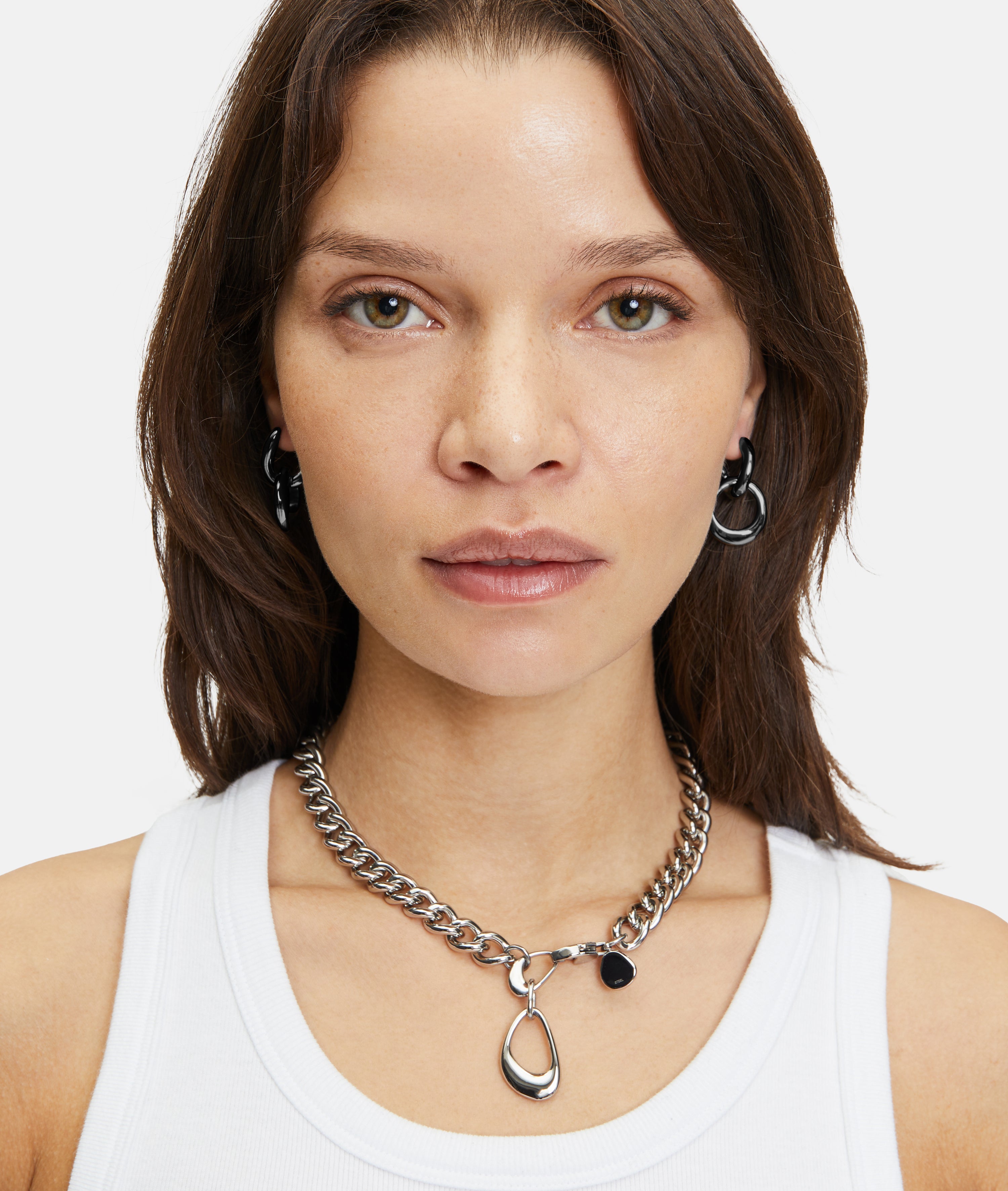 Frau mit glattem braunem Haar trägt ein weißes Tank-Top und Liebeskind Berlin-Schmuck, darunter The Organic Creole-Ohrringe (LIEBESKIND BERLIN Creole) und eine Halskette mit abnehmbarem Anhänger, mit Blick in die Kamera.