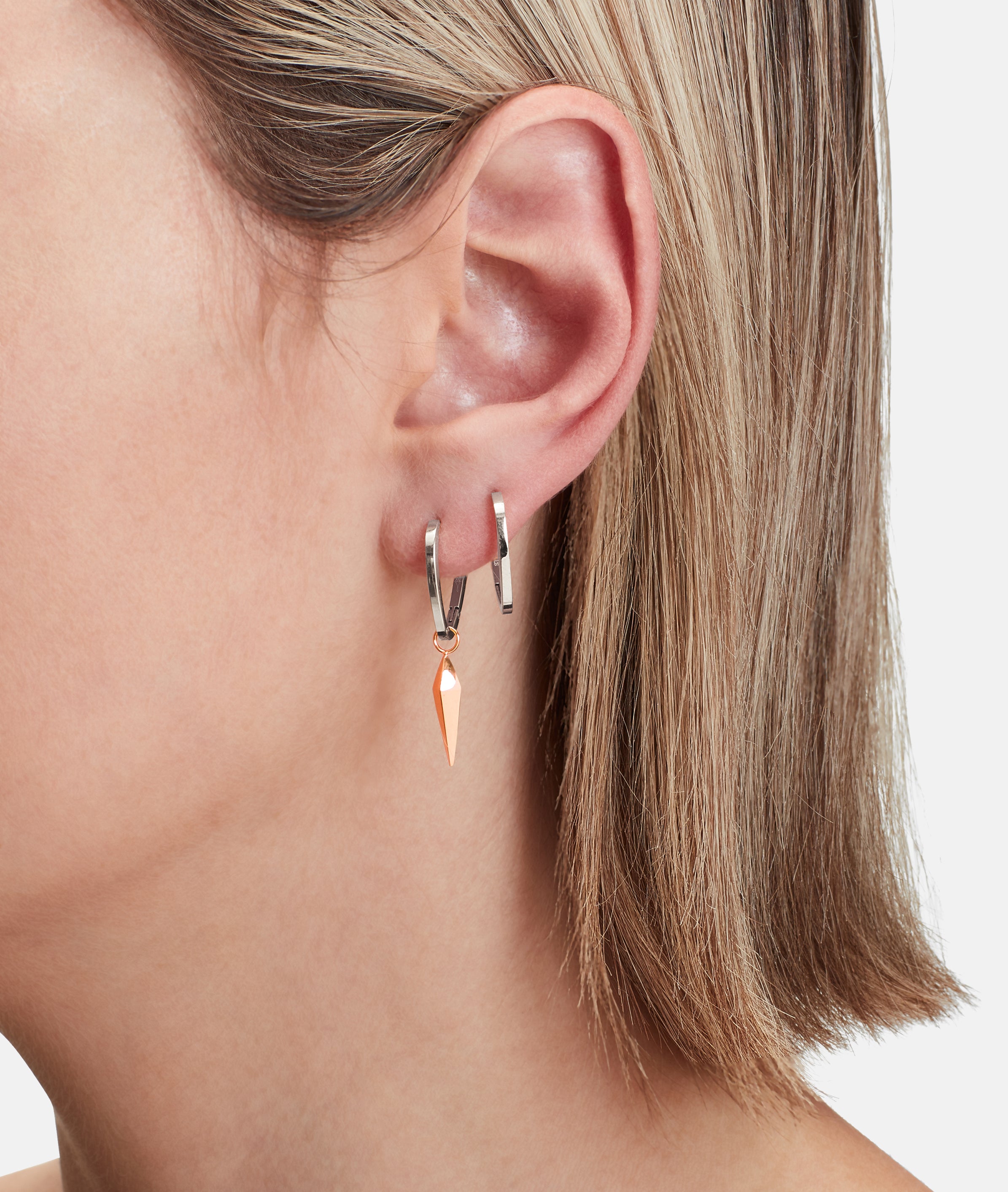 Nahaufnahme des Ohres einer Person mit zwei silbernen Creolen, eine davon mit einem spitzen orangefarbenen Anhänger. Entdecken Sie LIEBESKIND BERLIN Creole - The Pendulum, stilvoller Trendschmuck von Liebeskind Berlin.