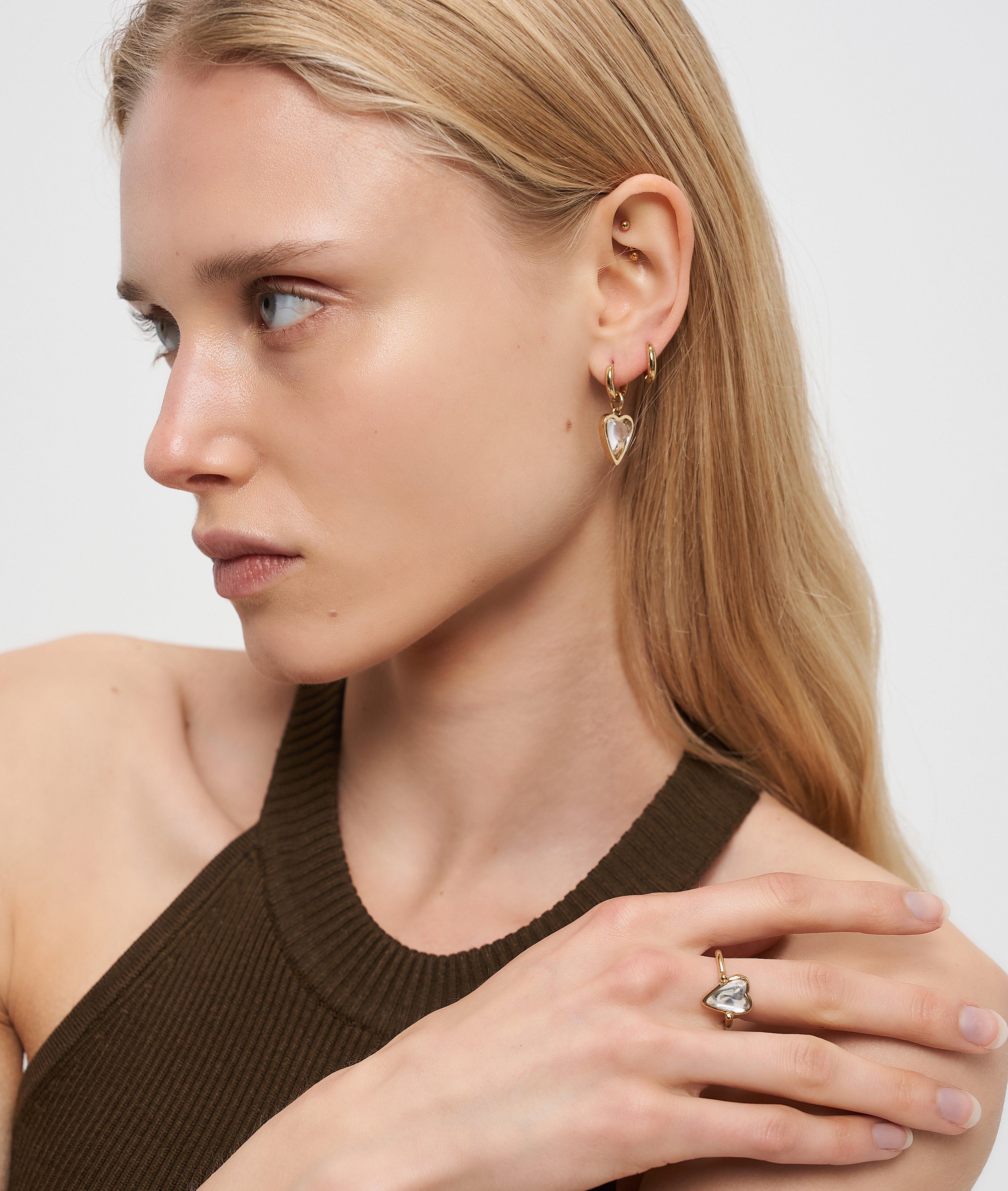 Frau mit blondem Haar trägt Liebeskind Berlin Creole - Glass Heart Ohrringe und einen passenden Ring, bekleidet mit einem braunen Neckholder-Top.