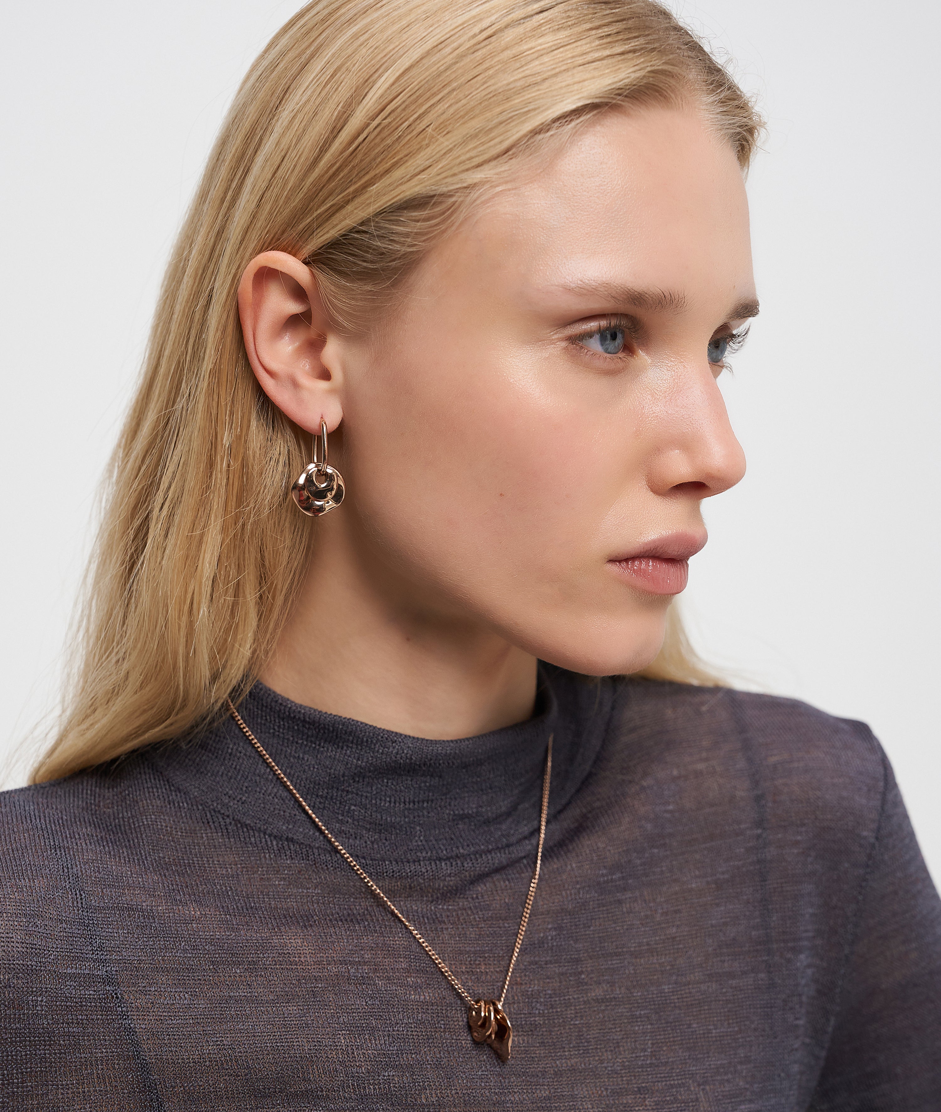 Frau mit langen blonden Haaren, die ein durchsichtiges dunkles Oberteil, Ohrringe und eine Liebeskind Berlin Halskette - Organic Shapes trägt und zur Seite schaut.