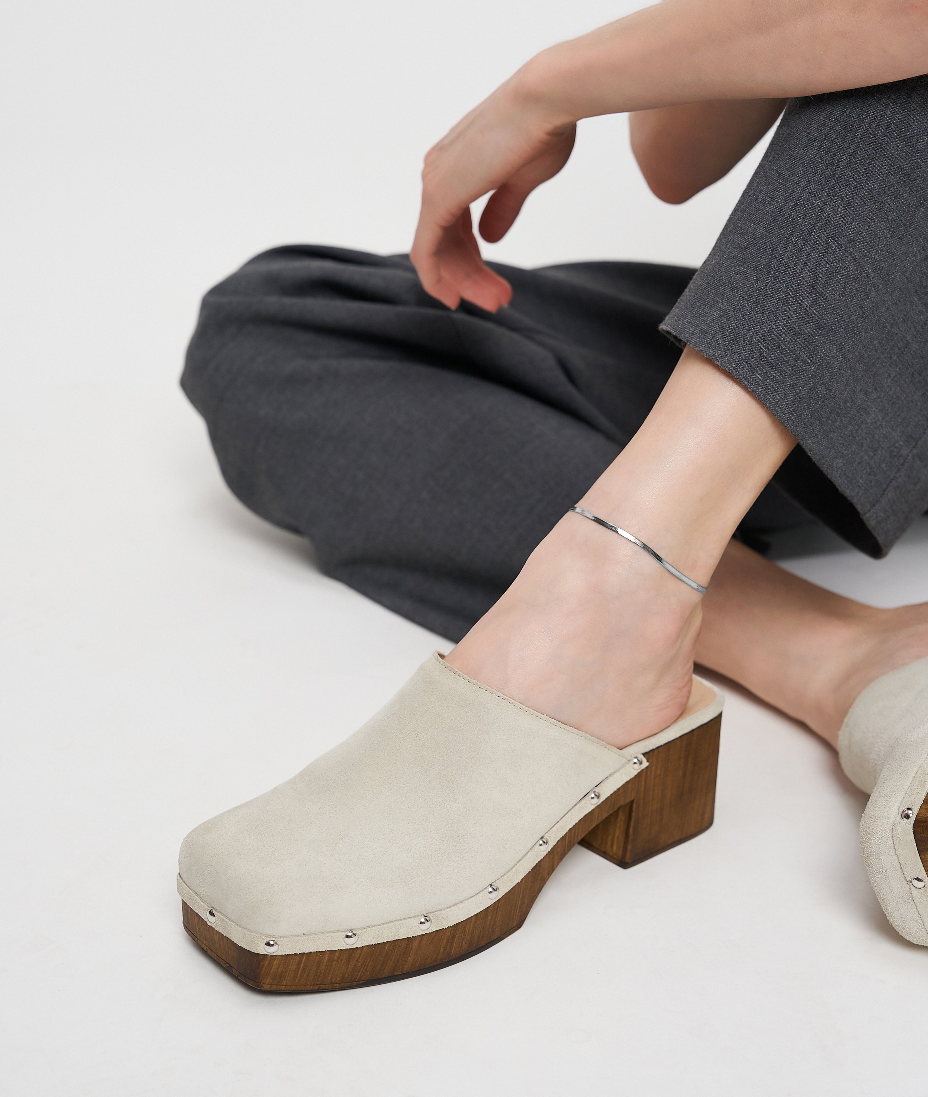 Auf einem weißen Hintergrund sitzt eine Person in beigen Wildlederclogs und grauer Hose und präsentiert die Liebeskind Berlin Fußkette - The Sleek.