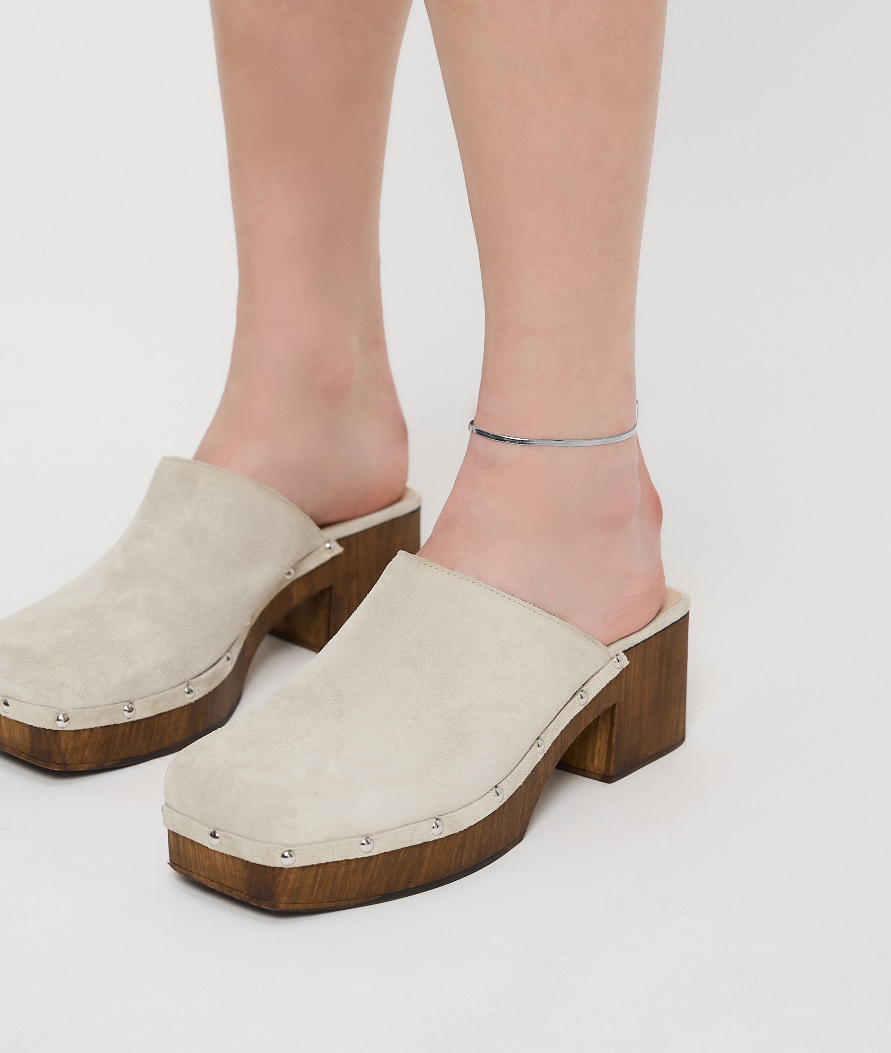 Person in beigen Plateau-Clogs mit Holzabsatz und einer Liebeskind Berlin Fußkette - The Sleek von Liebeskind Berlin auf weißem Hintergrund.