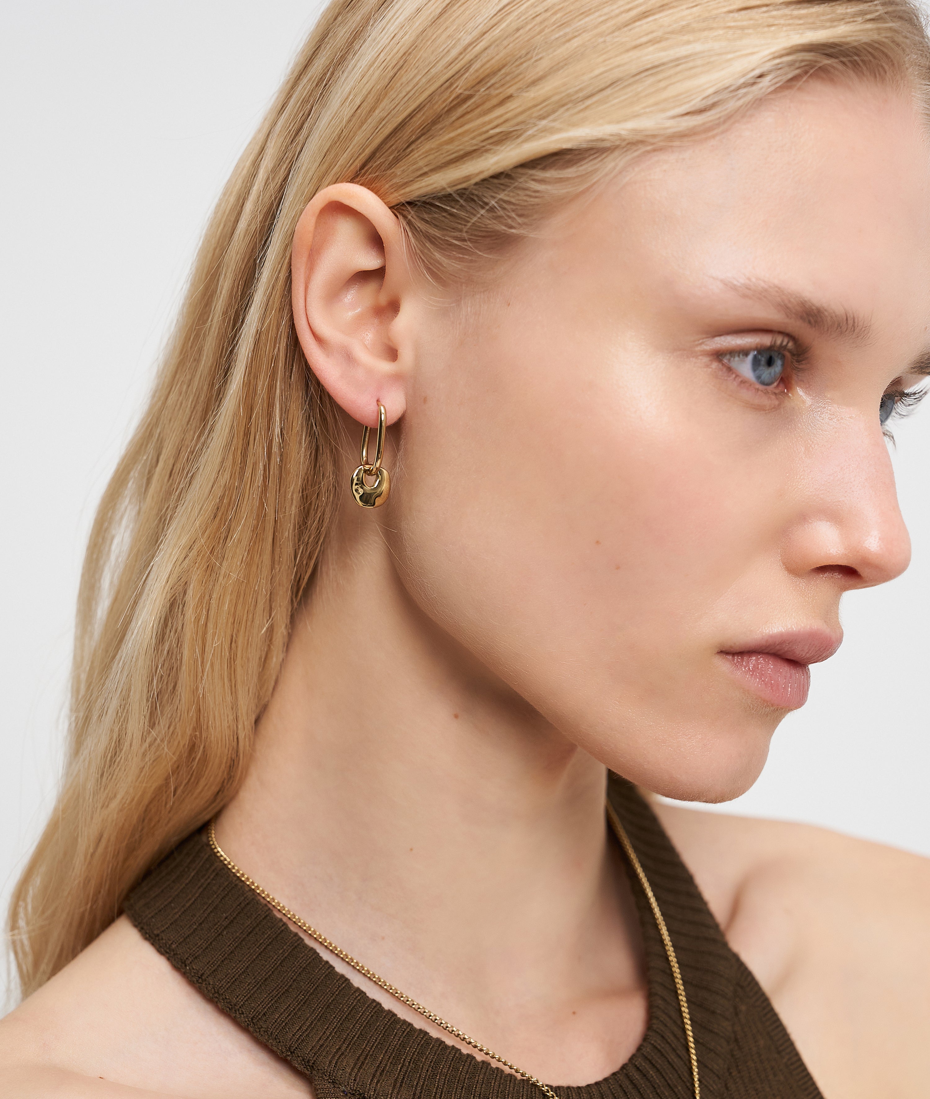 Frau mit langen blonden Haaren trägt Liebeskind Berlin Ohrring - Organic Shapes, goldene Creolen von Liebeskind Berlin, gepaart mit einem braunen Neckholder-Top, von rechts.
