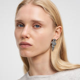 Eine Frau mit glattem blondem Haar trägt den Liebeskind Berlin Ohrring - Organic Shapes und einen hellgrauen Pullover und schaut in die Kamera.