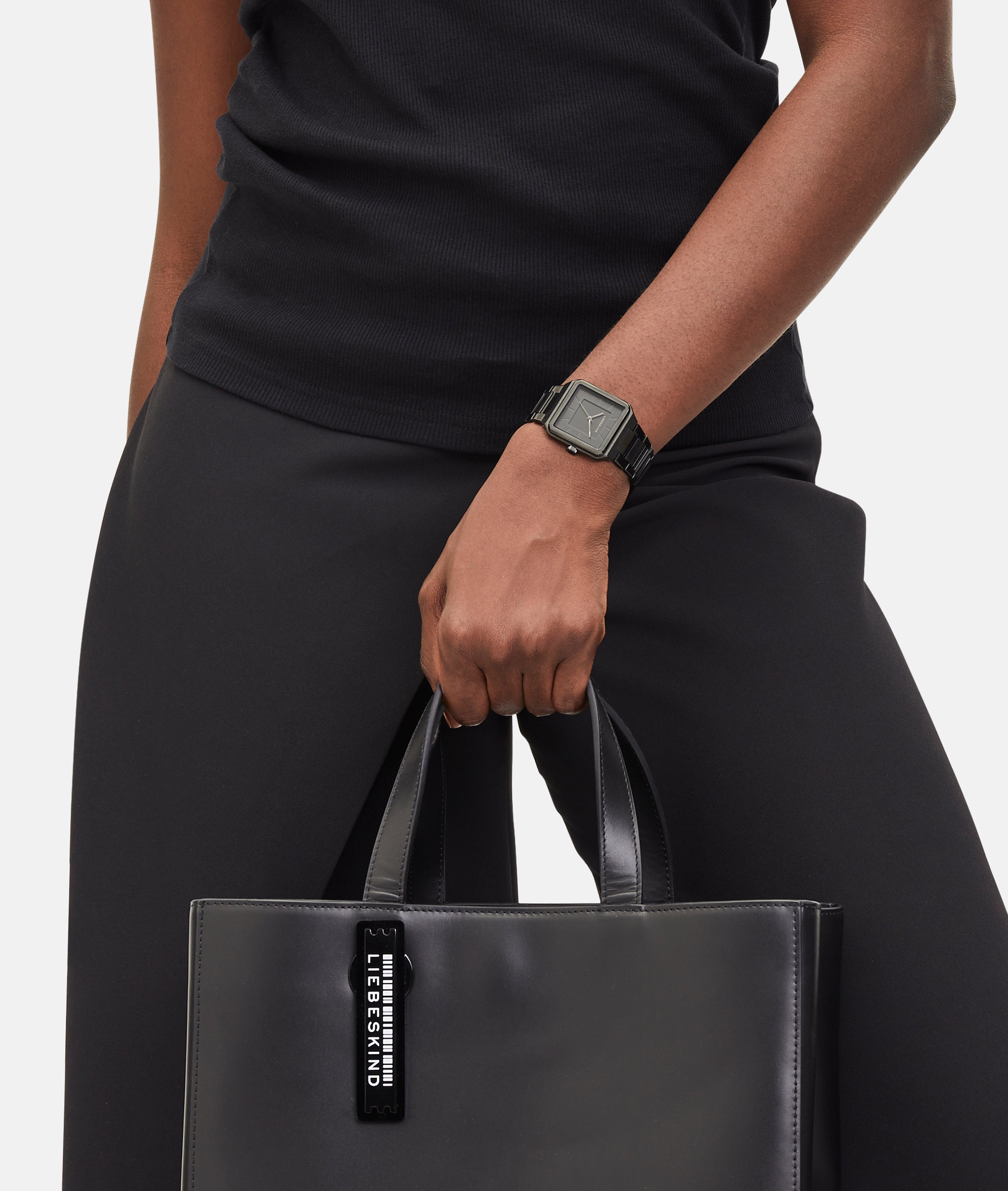 Eine Person in Schwarz, die ein ärmelloses Oberteil trägt und eine schwarze Handtasche in der Hand hält, trägt die elegante Liebeskind Berlin Armbanduhr - The Square Minimalist.