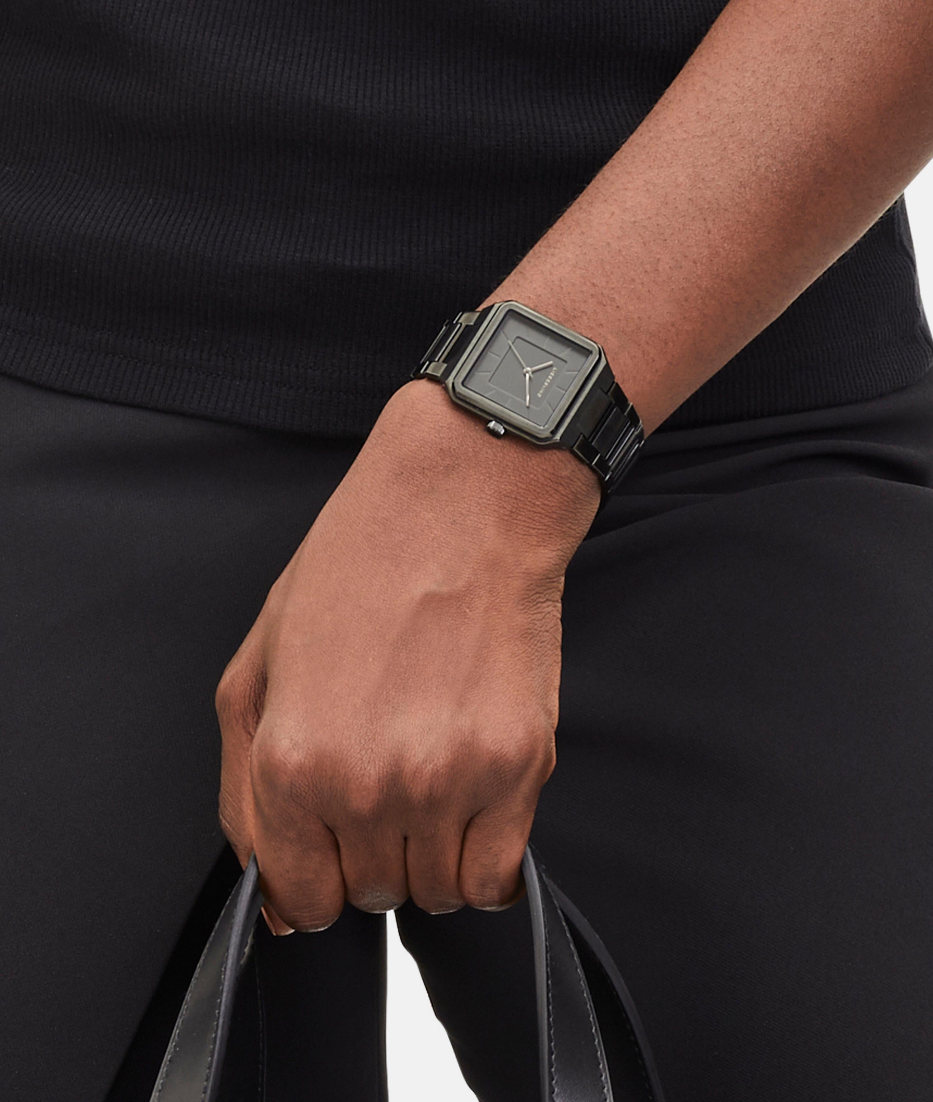 Eine Person in schlichter schwarzer Kleidung hält eine schwarze Tasche und trägt die LIEBESKIND BERLIN Armbanduhr - The Square Minimalist von Liebeskind Berlin.