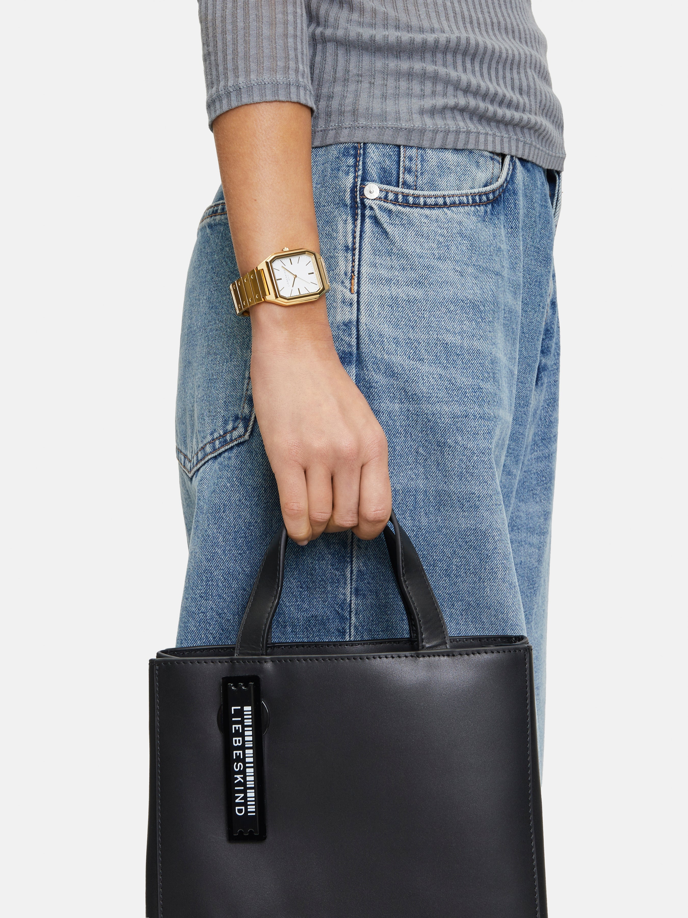Eine Person in Jeans und einem gerippten grauen Oberteil trägt eine schwarze Handtasche und präsentiert die Liebeskind Berlin Armbanduhr - The Basic Staple an ihrem Handgelenk.