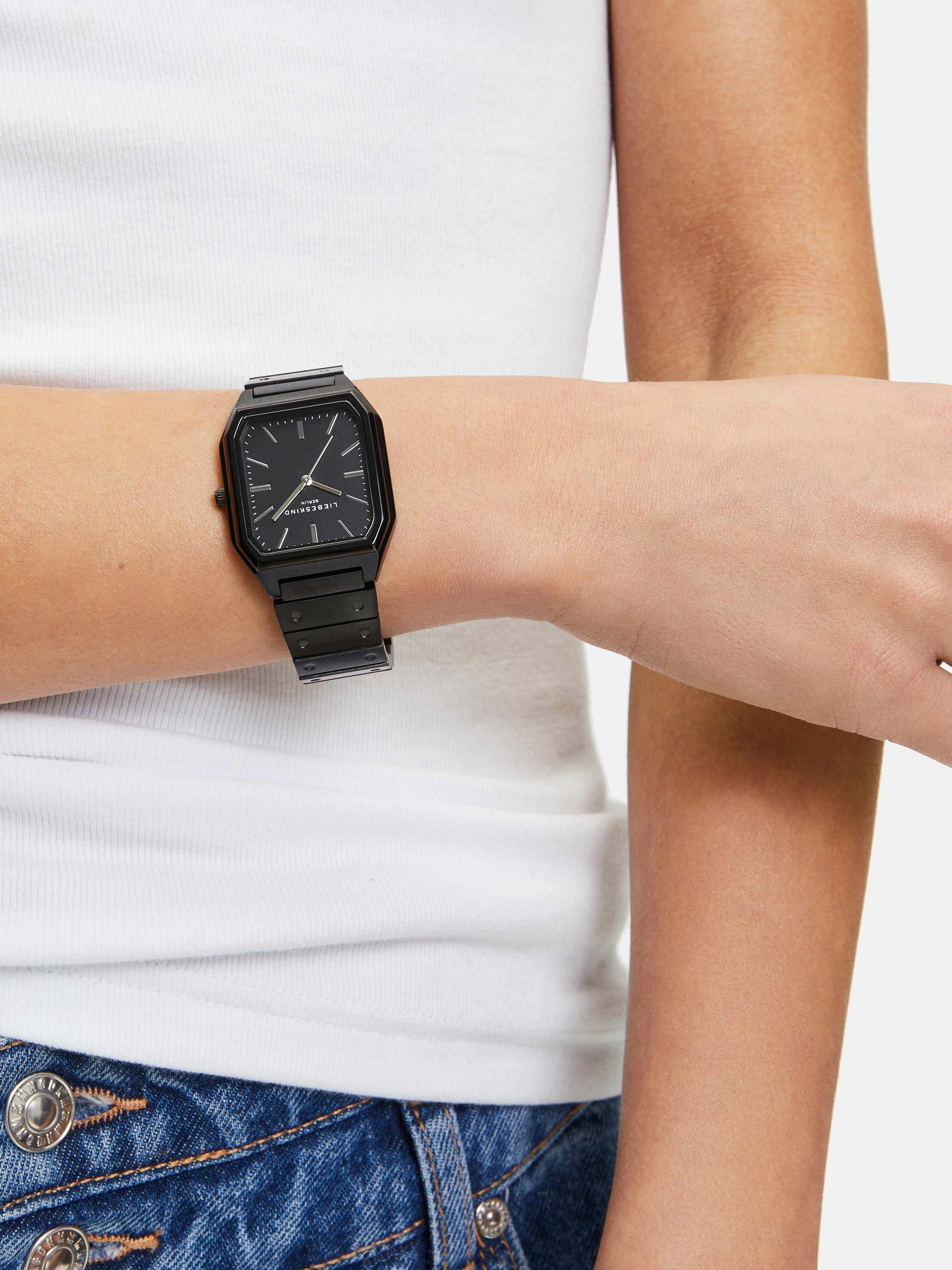 Eine Person trägt eine Liebeskind Berlin Armbanduhr - The Basic Staple, ein weißes Tanktop und eine blaue Jeans mit silbernen Knöpfen.