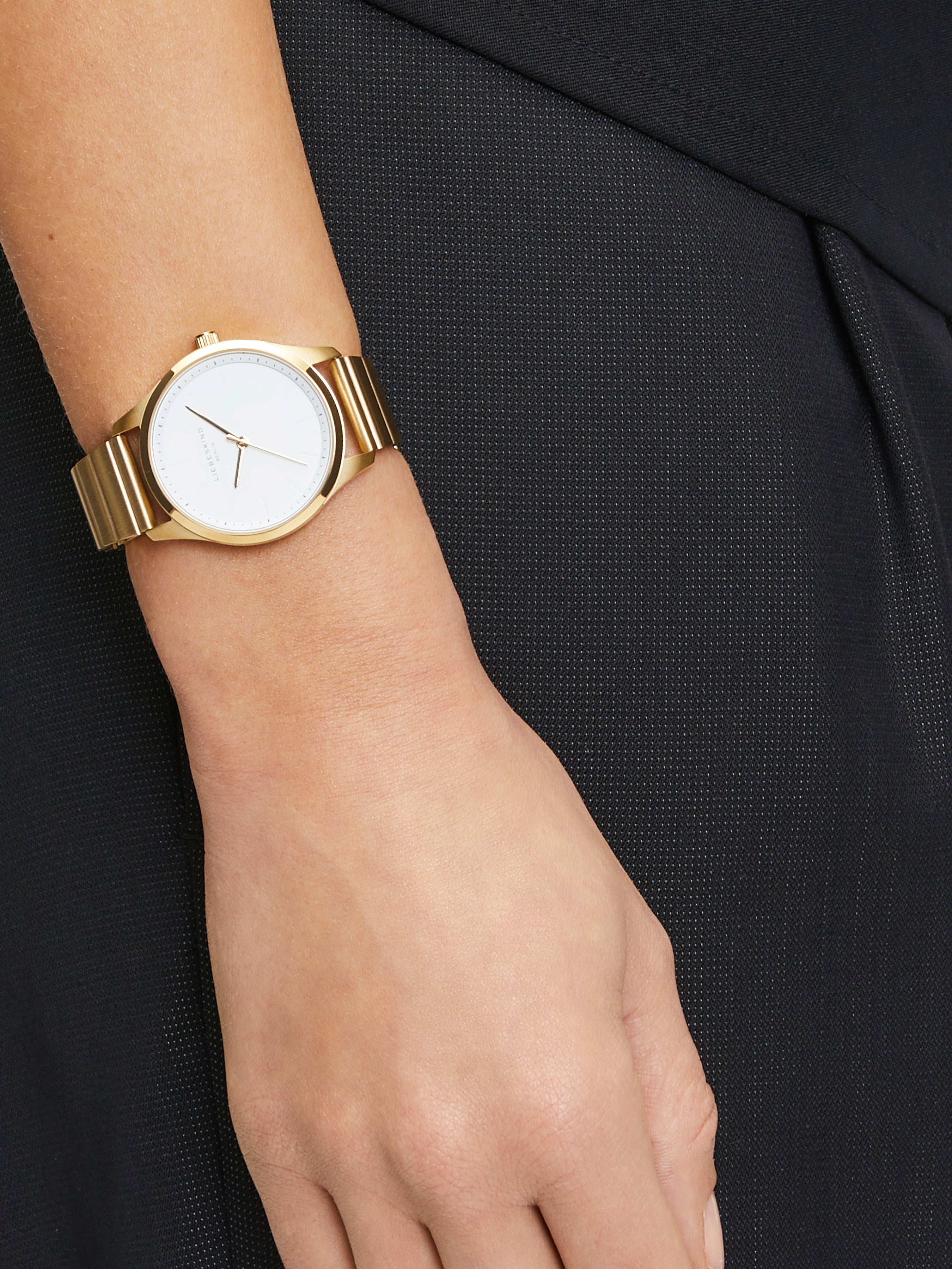 Die LIEBESKIND BERLIN Armbanduhr - The Round Staple überzeugt mit modernem Design, goldfarbenem Band und weißem Zifferblatt und setzt stilvolle Akzente zu schwarzer, strukturierter Kleidung.