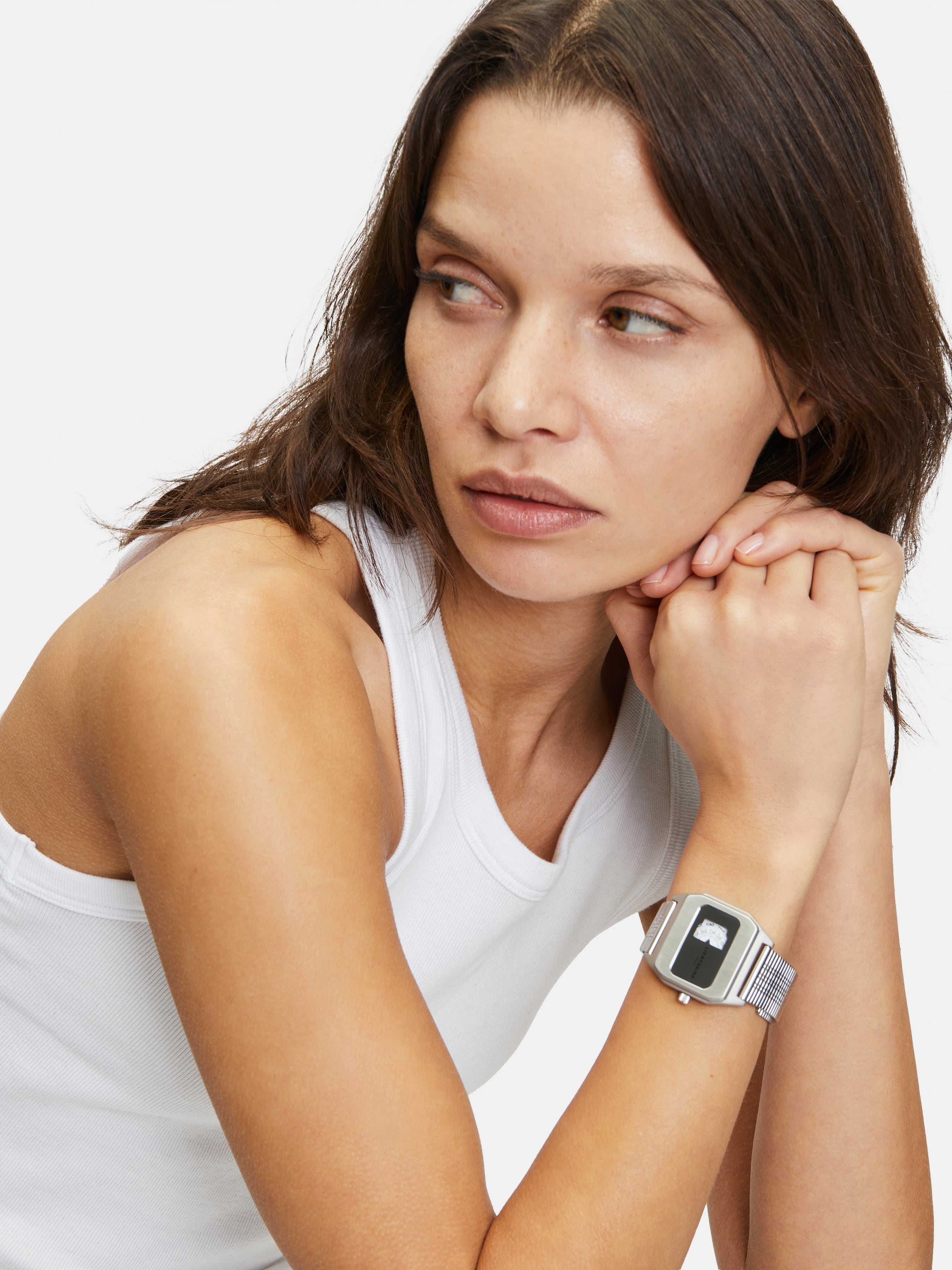 Eine Frau in einem weißen Tank-Top schaut zur Seite und stützt ihr Kinn auf die Hände. Sie trägt die Liebeskind Berlin Armbanduhr - Vinyl Vision.