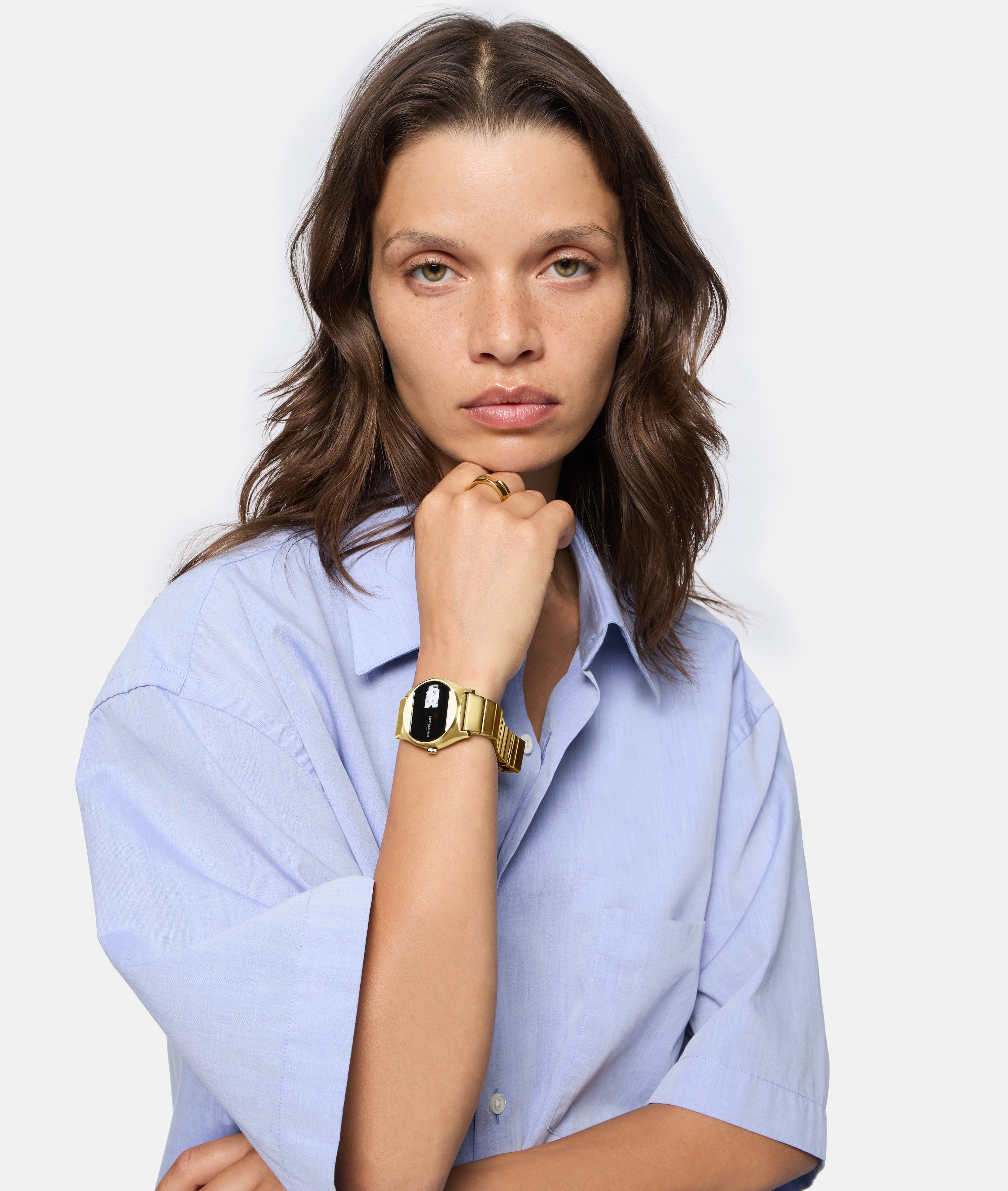 Eine Frau mit hellblauem Hemd trägt die LIEBESKIND BERLIN Armbanduhr - Round Vinyl Vision gold von Liebeskind Berlin mit verspiegeltem Ziffernblatt, während sie vor einem schlichten Hintergrund ihr Kinn auf ihre Hand stützt.
