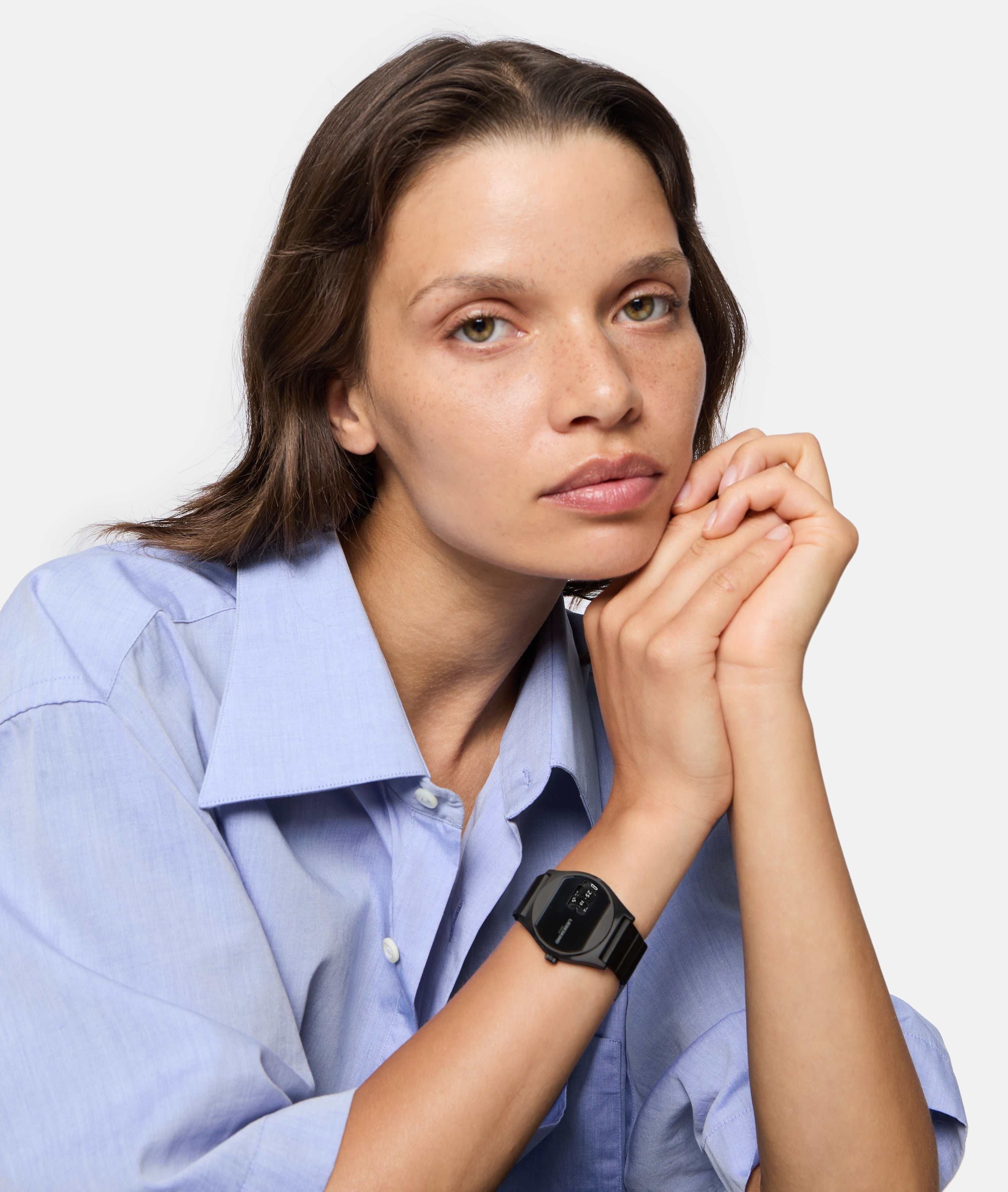 Eine Frau in einem hellblauen Hemd mit gefalteten Händen trägt die Liebeskind Berlin Armbanduhr - Round Vinyl Vision und schaut in die Kamera.