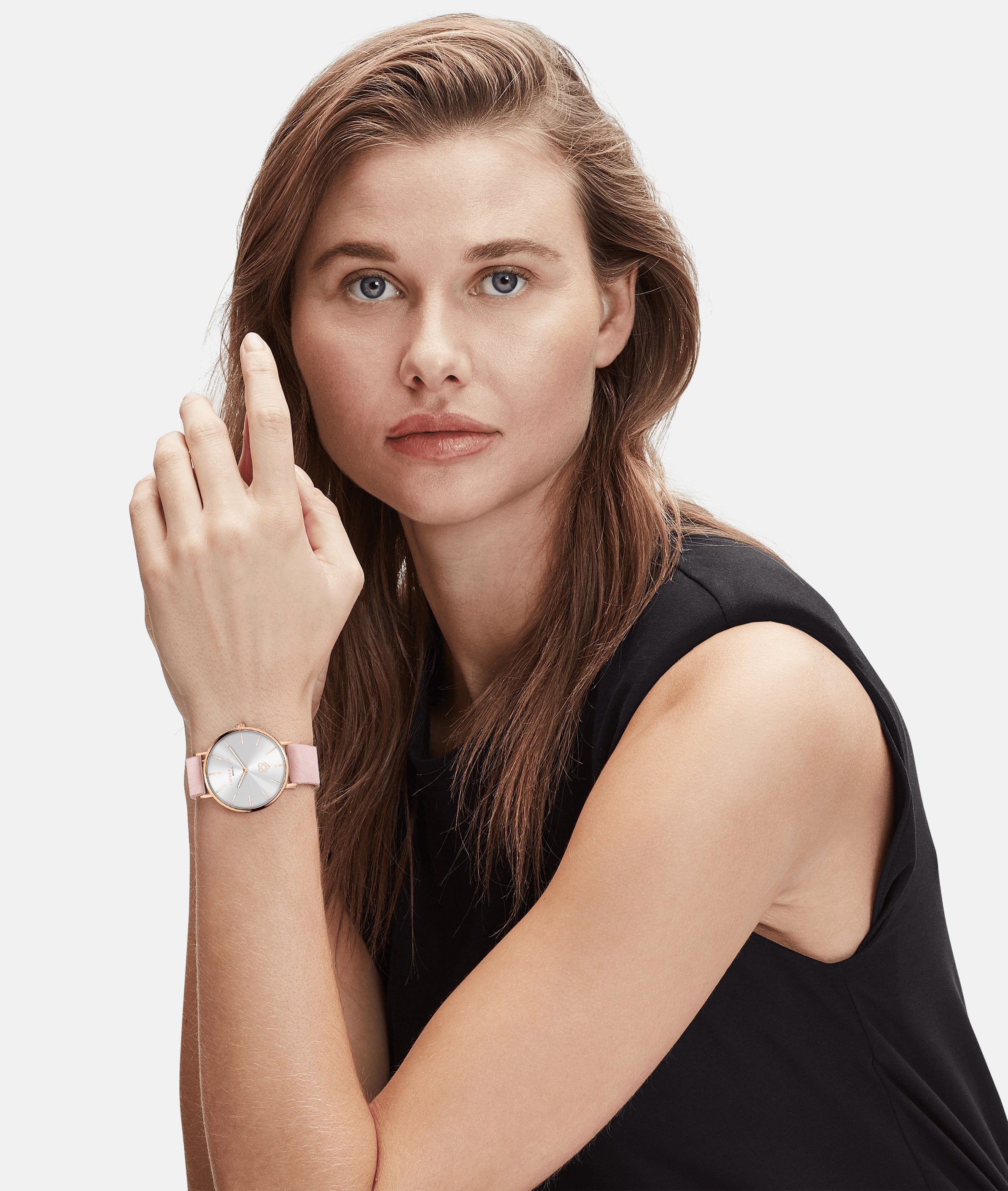 Frau mit hellbraunem Haar, die ein ärmelloses schwarzes Oberteil und eine rosafarbene LIEBESKIND BERLIN Armbanduhr - Keep Loving im skandinavischen Design von Liebeskind Berlin trägt, schaut in die Kamera.
