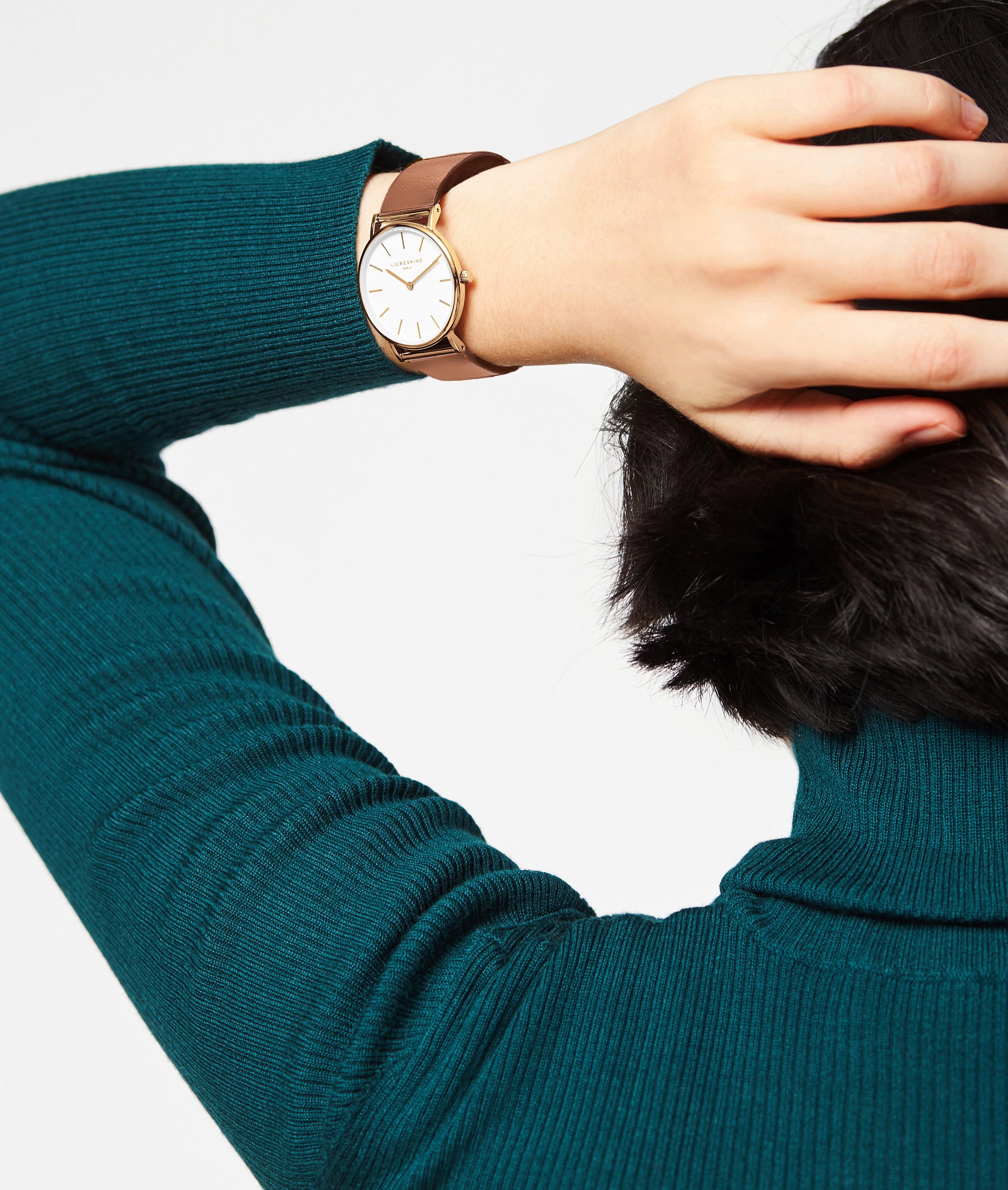 Eine Person im türkisfarbenen Pullover trägt am Handgelenk die LIEBESKIND BERLIN Armbanduhr - Das moderne Must-Have von Liebeskind Berlin mit Echtlederarmband und goldenem Gehäuse.