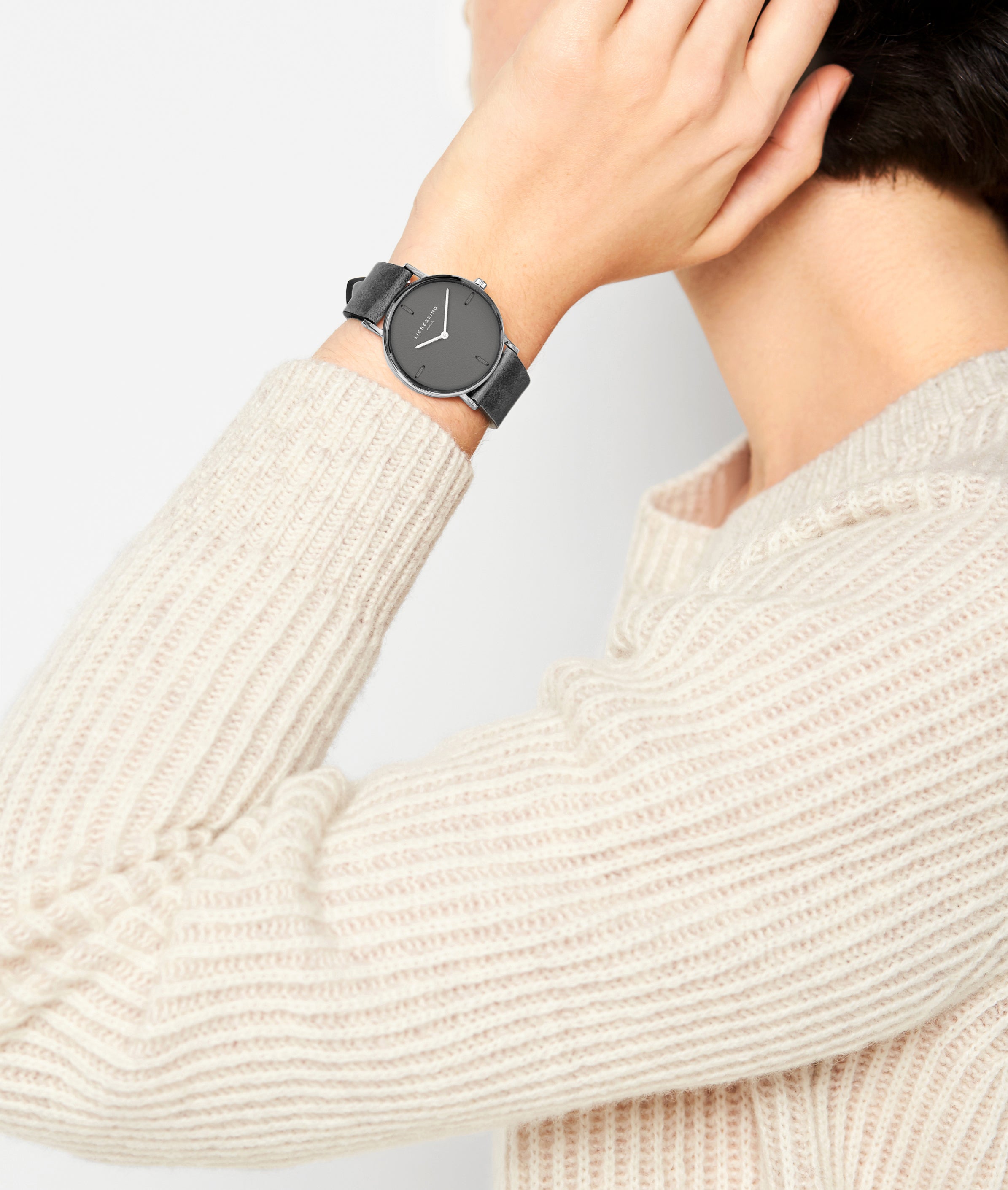 Die Person trägt einen cremefarbenen Strickpullover und eine Liebeskind Berlin Armbanduhr - The Leather One, wobei sie die Hand an ihr Ohr hält.