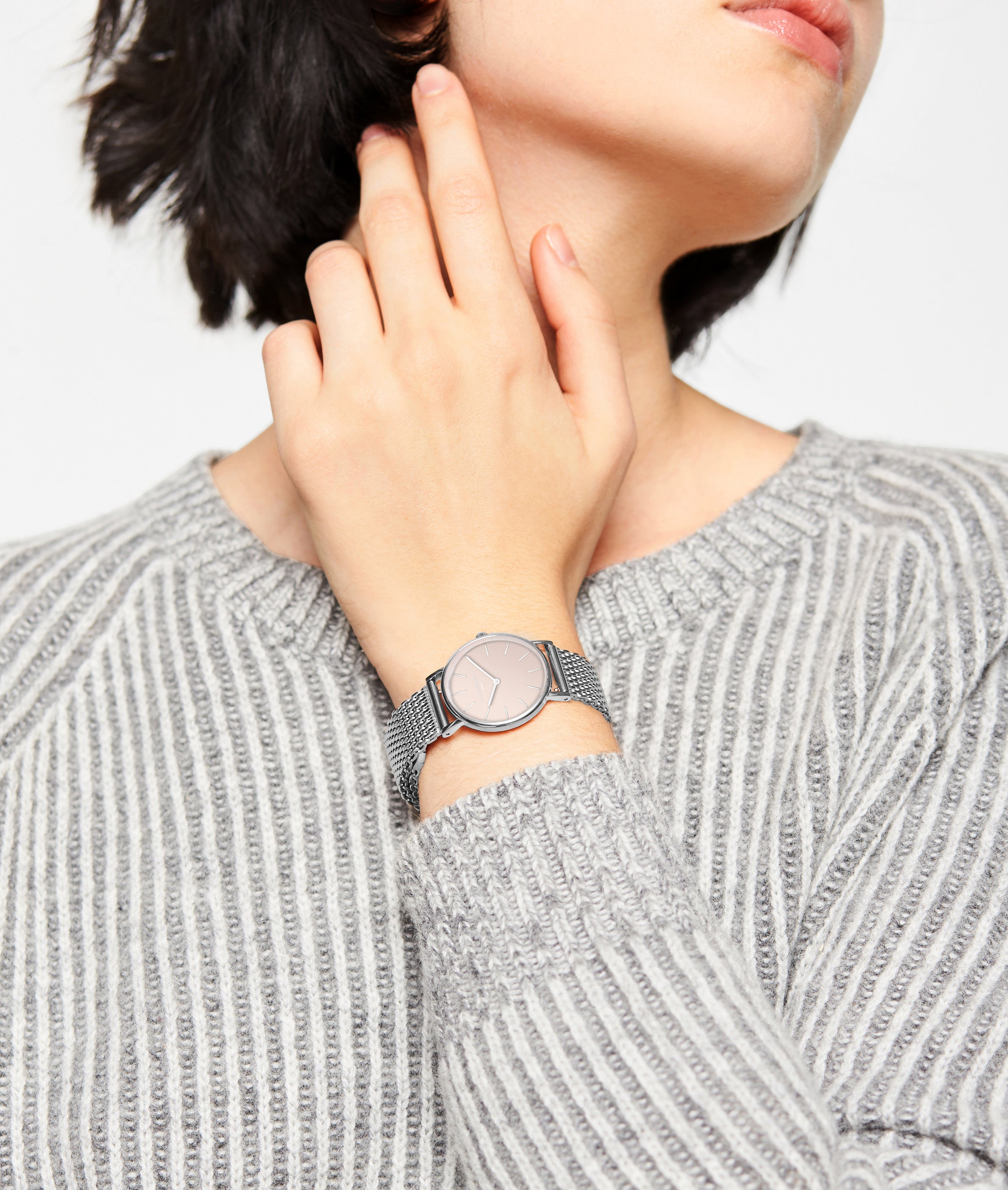 Eine Person in einem grau gestreiften Pullover berührt ihren Hals und trägt die LIEBESKIND BERLIN Armbanduhr - Color Up von Liebeskind Berlin, eine elegante Edelstahluhr mit Netzband und minimalistischem skandinavischem Design.