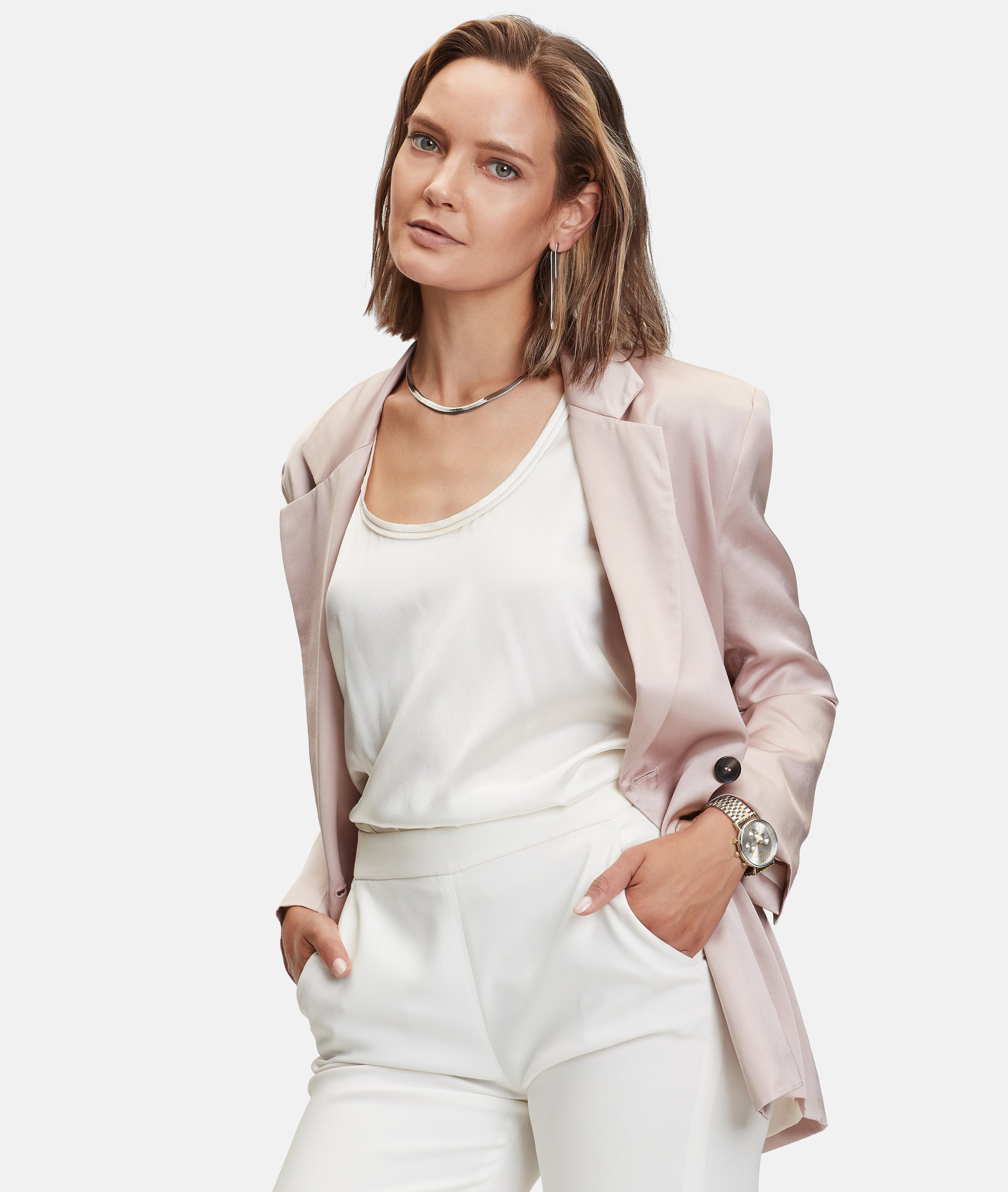 Frau in hellrosa Blazer und weißem Outfit, Hände in den Taschen, blickt selbstbewusst in die Kamera, während sie die stilvolle LIEBESKIND BERLIN Armbanduhr - Modern Chronograph von Liebeskind Berlin mit einem eleganten Edelstahlarmband trägt.