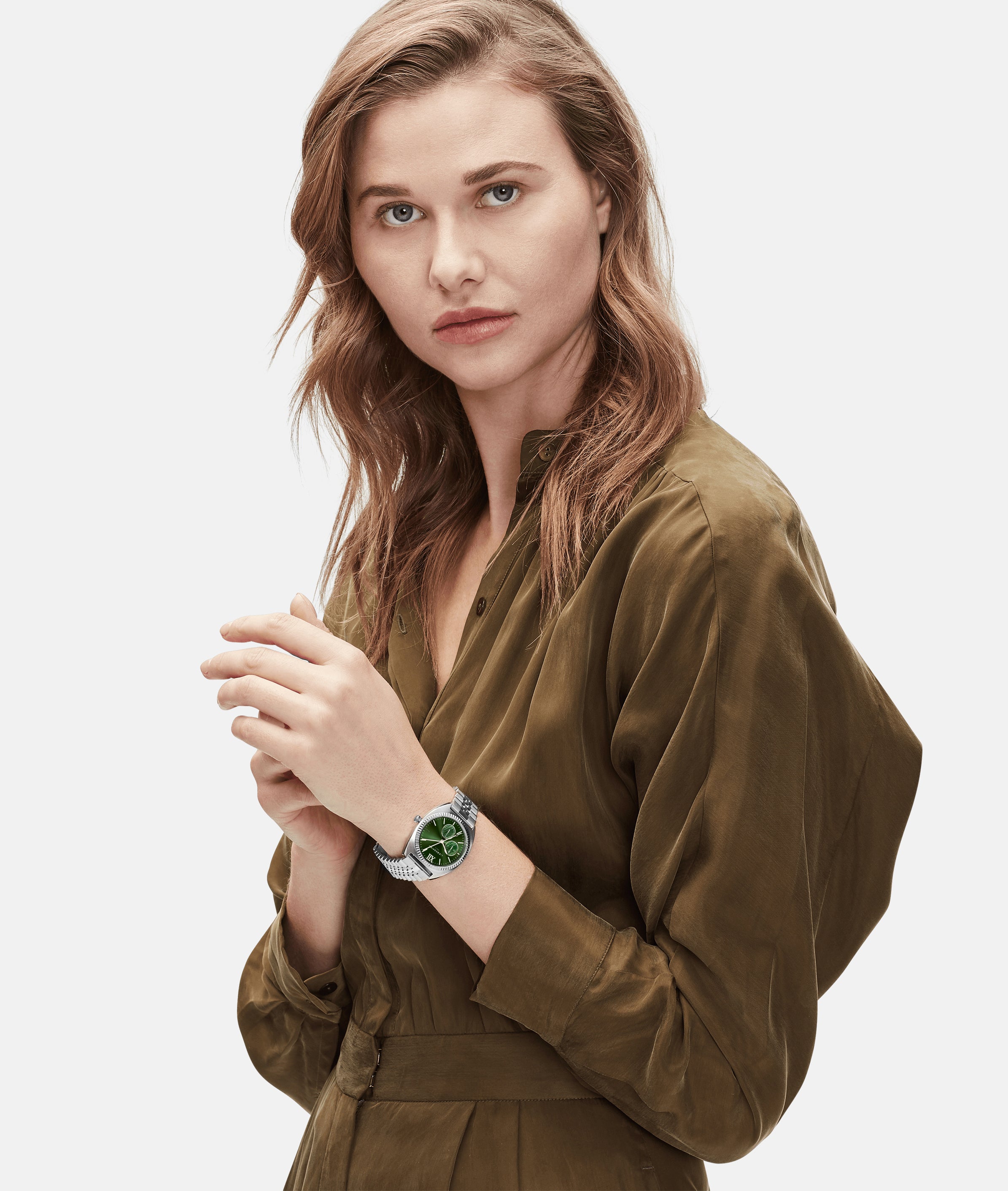 Eine Frau in einem olivgrünen Kleid trägt die LIEBESKIND BERLIN Armbanduhr - The Colorful Multifunction von Liebeskind Berlin mit grünem Zifferblatt und elegantem Edelstahlarmband vor einem weißen Hintergrund.