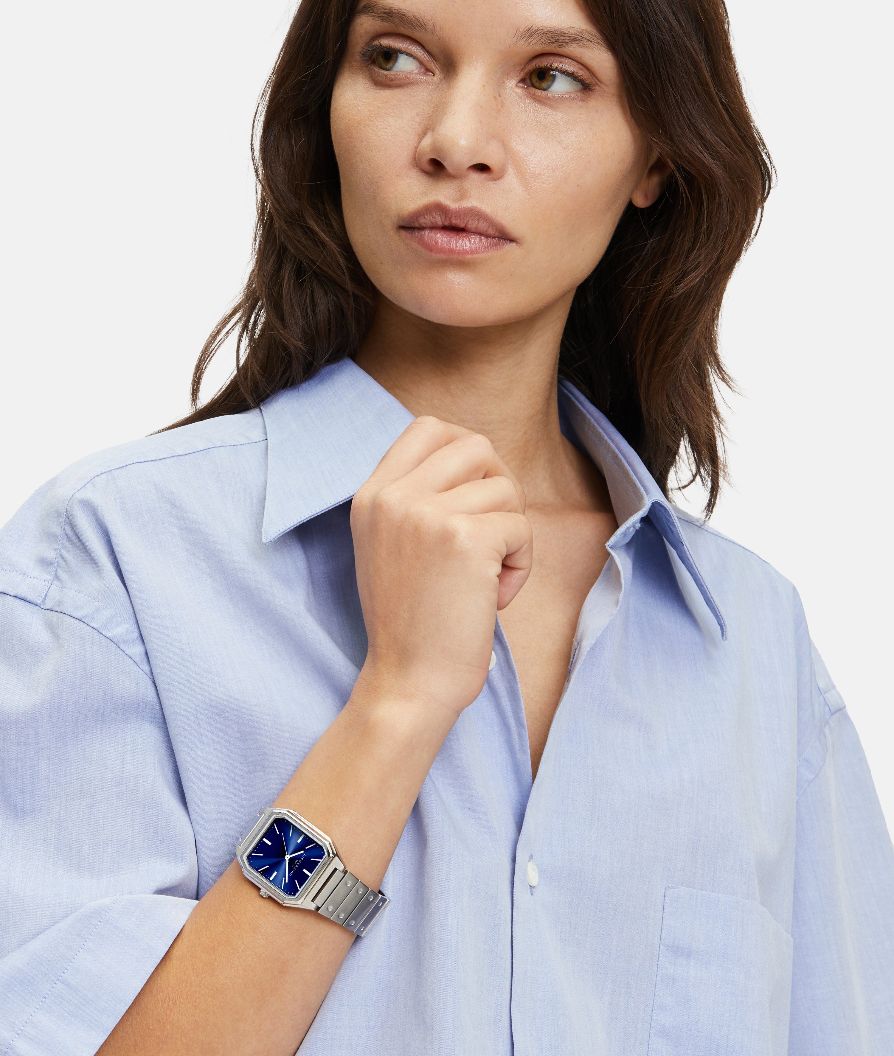 Frau in hellblauem Hemd trägt die Liebeskind Berlin Armbanduhr - The Modern Staple, eine elegante Quarzuhr mit blauem, quadratischem Zifferblatt, mit Blick zur Seite.