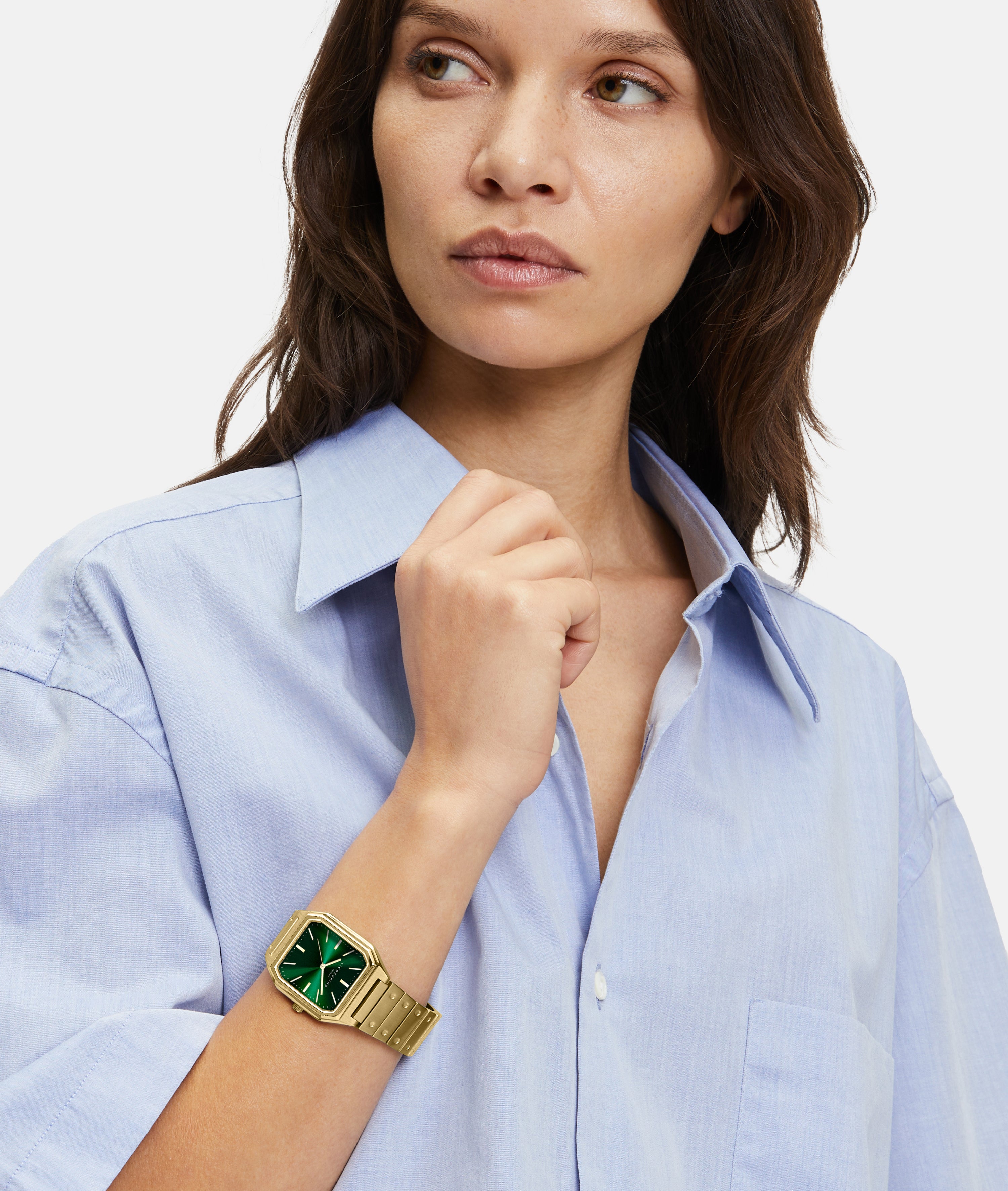 Frau in hellblauem Hemd trägt die Liebeskind Berlin Armbanduhr - The Modern Staple mit rechteckigem Gehäuse und grünem Zifferblatt, Blick zur Seite.