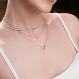 Frau trägt eine Ania Haie Halskette - Flache Schlangenkette aus 925 Sterling Silber, gestylt im Dreilagen-Look mit Kreuz- und Herzanhängern über einem weißen Top.