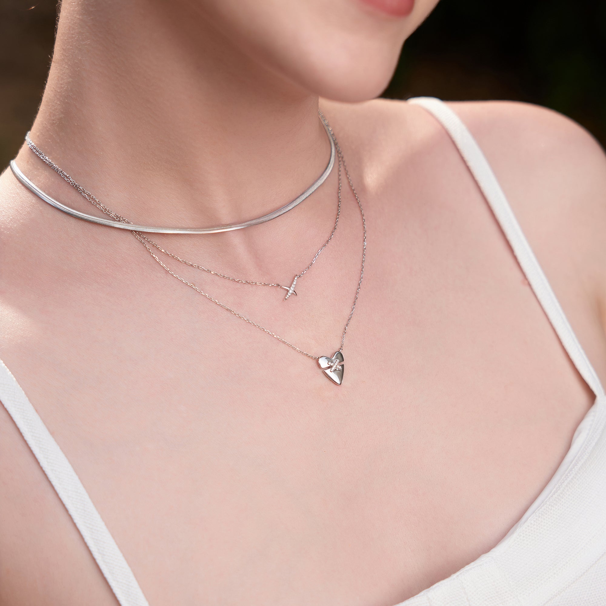 Frau trägt eine Ania Haie Halskette - Flache Schlangenkette aus 925 Sterling Silber, gestylt im Dreilagen-Look mit Kreuz- und Herzanhängern über einem weißen Top.