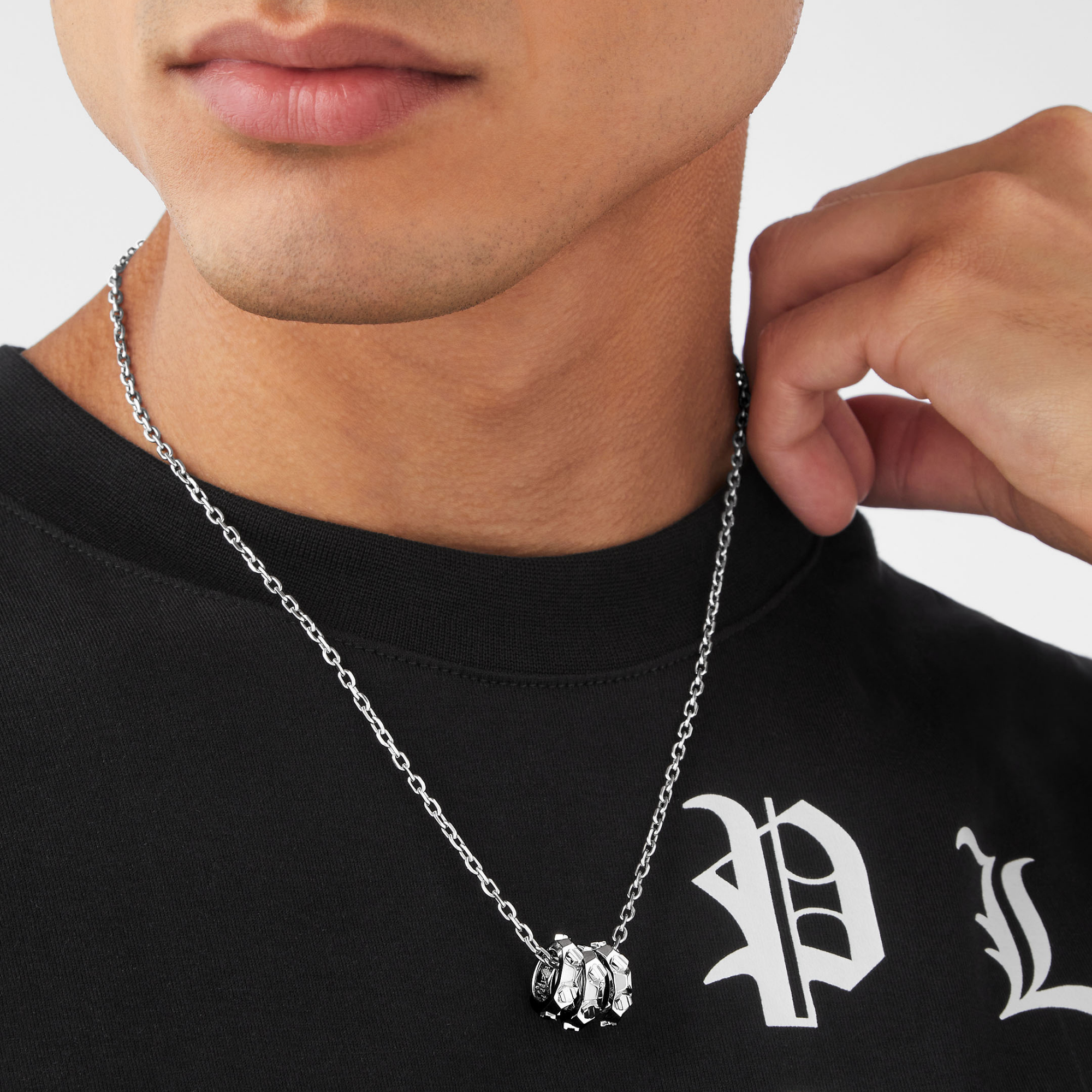 Eine Person trägt die Philipp Plein Halskette - PLEIN CUFF, eine silberne Kette mit drei auffälligen Ringen von Philipp Plein, die als Statement-Schmuck gestylt ist, gepaart mit einem schwarzen Hemd mit weißen gotischen Buchstaben.