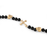 Das Philipp Plein Armband - PP WHISPER von Philipp Plein ist mit schwarzen Perlen, goldenen Totenköpfen und einem goldenen Kreuz verziert und hebt Schwarz und Gold vor einem weißen Hintergrund hervor.