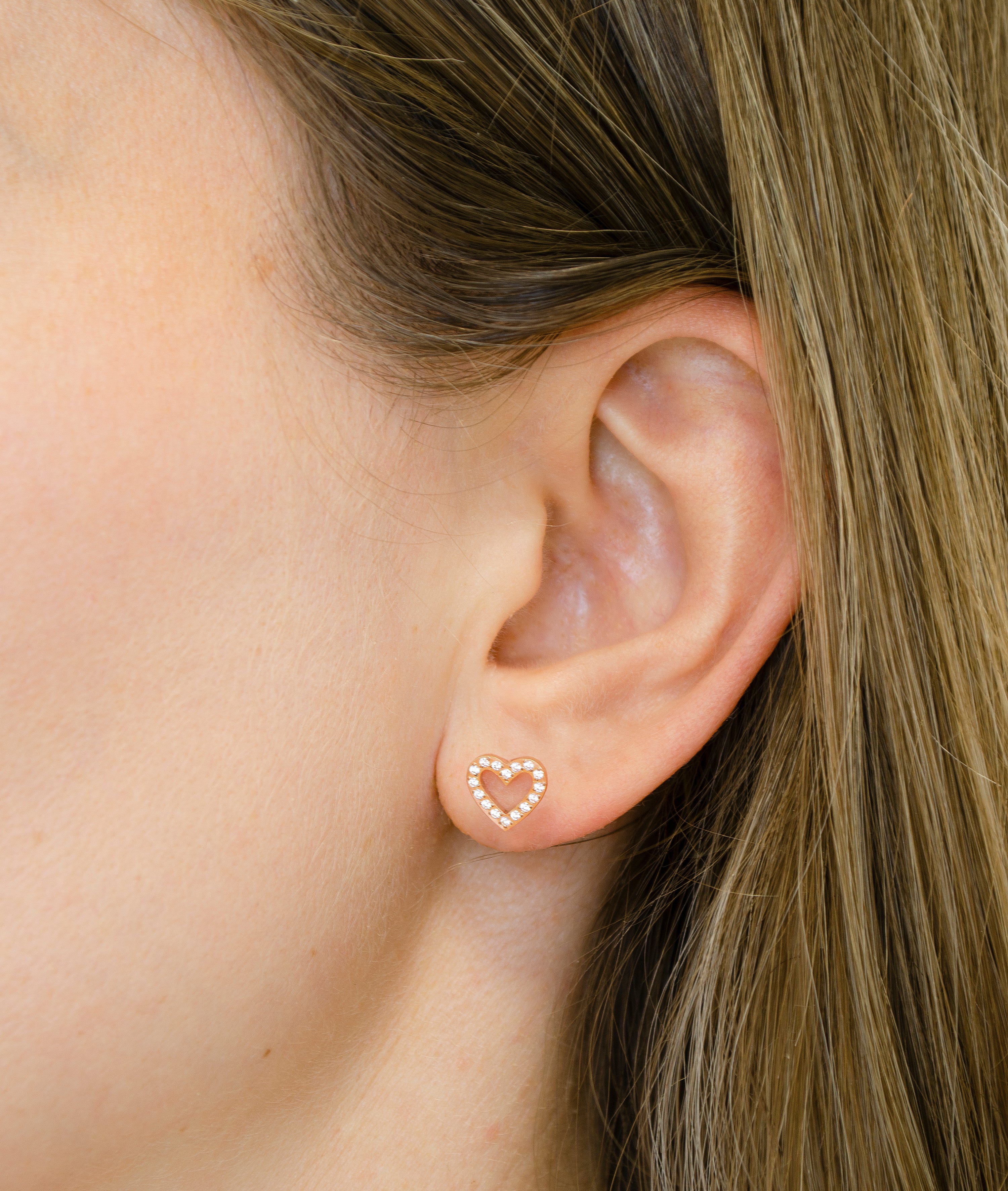 Nahaufnahme eines Ohrs mit hellbraunem Haar, das einen Tamaris Ohrstecker von Tamaris trägt, einen goldenen herzförmigen Ohrstecker, der mit kleinen Kristallen besetzt ist.