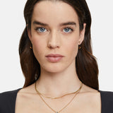Hellhäutige Frau mit blauen Augen und langen braunen Haaren, nach vorne blickend, trägt die goldenen Ohrringe The 2 in 1 Creole von Tamaris und zarte goldene Halsketten.