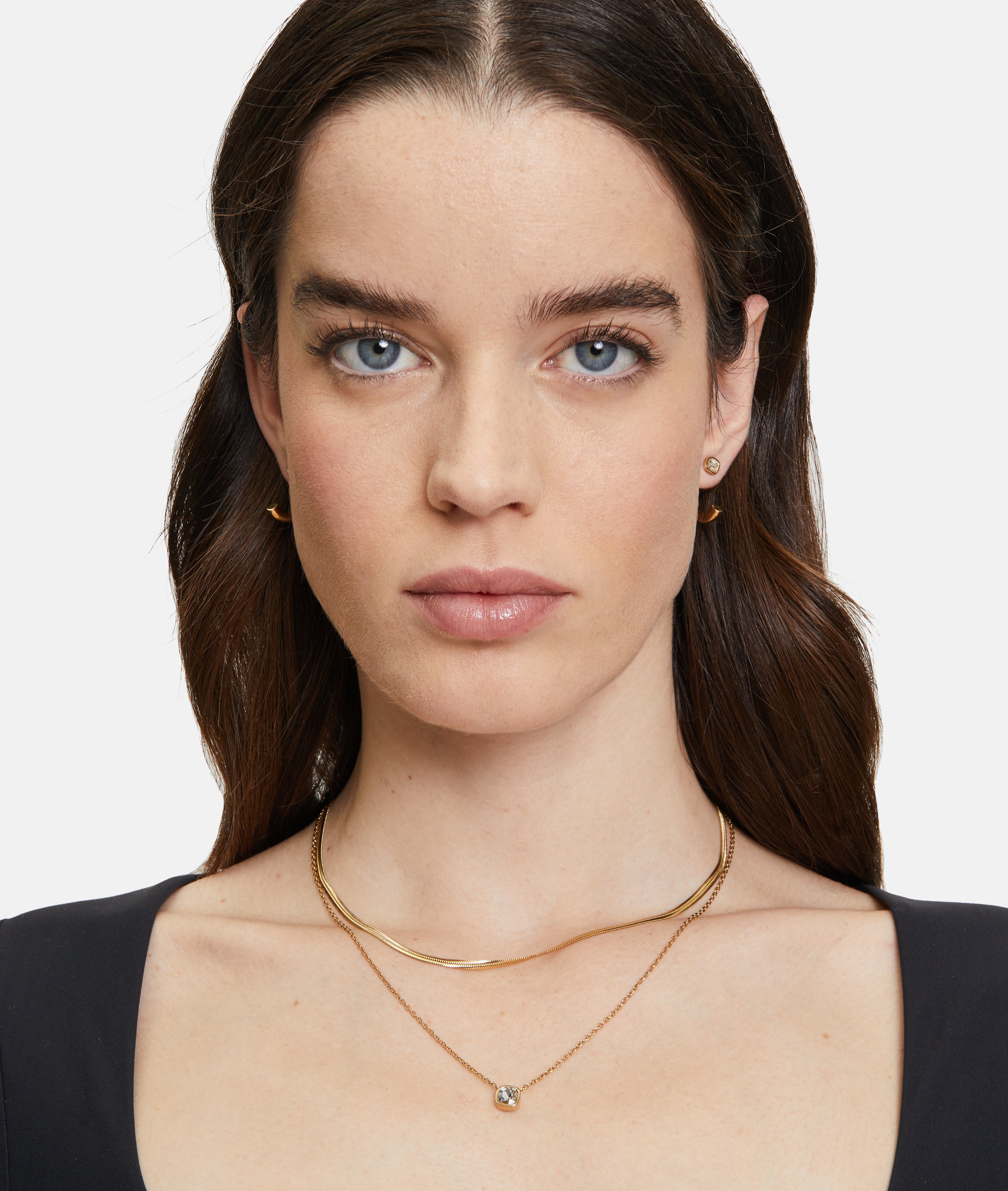 Hellhäutige Frau mit blauen Augen und langen braunen Haaren, nach vorne blickend, trägt die goldenen Ohrringe The 2 in 1 Creole von Tamaris und zarte goldene Halsketten.
