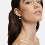 Eine Frau mit langen braunen Haaren trägt goldene, geblümte Ohrringe und eine Tamaris Halskette - The Flower Pearl, die leicht zur Seite zeigt.