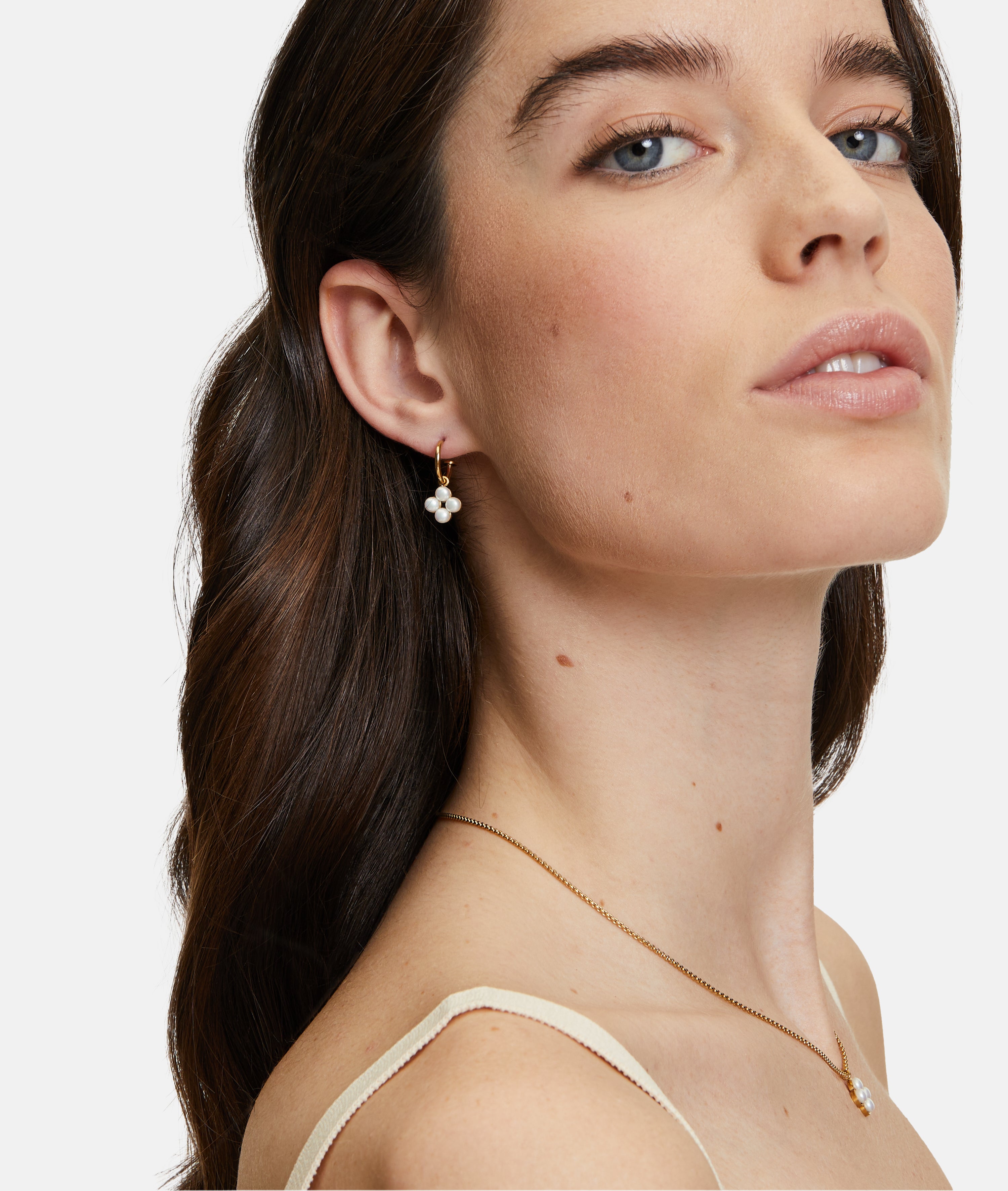 Eine Frau mit langen braunen Haaren trägt goldene, geblümte Ohrringe und eine Tamaris Halskette - The Flower Pearl, die leicht zur Seite zeigt.