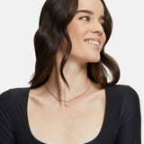 Eine Frau mit gewelltem braunem Haar lächelt, sie trägt ein schwarzes Oberteil und die Tamaris Halskette - The Flower Pearl von Tamaris mit einem zarten Glasperlen-Anhänger.