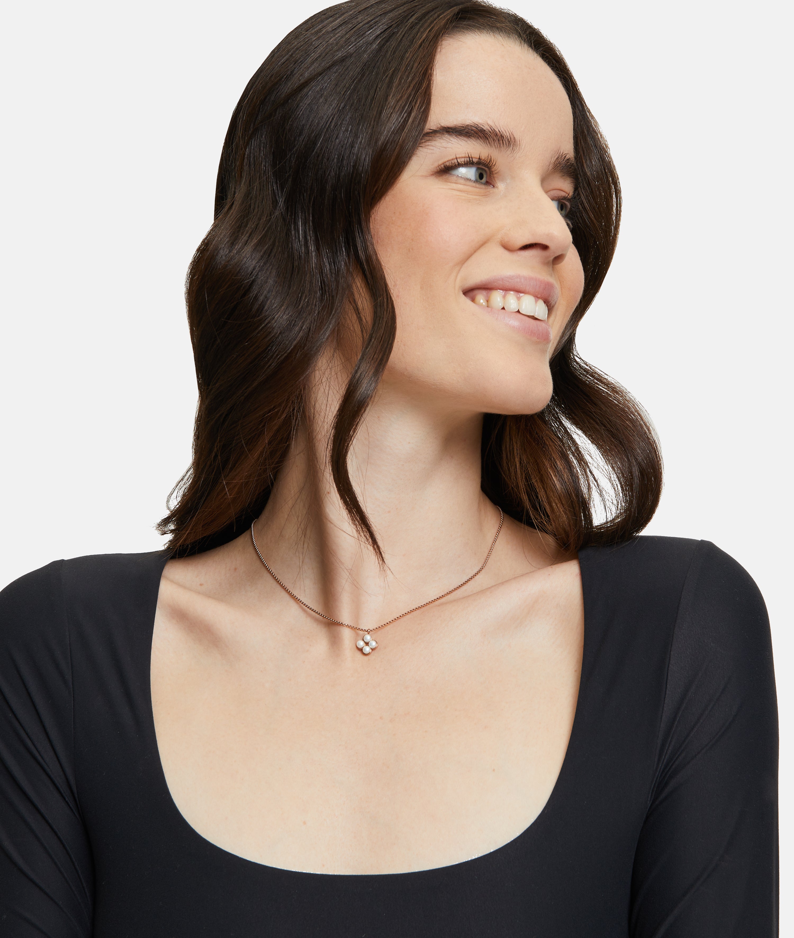 Eine Frau mit gewelltem braunem Haar lächelt, sie trägt ein schwarzes Oberteil und die Tamaris Halskette - The Flower Pearl von Tamaris mit einem zarten Glasperlen-Anhänger.