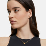 Eine Frau mit langen braunen Haaren trägt ein schwarzes Top, eine goldene Herzkette und den Tamaris Ohrstecker - The T-Pattern Heart von Tamaris - perfekt als Geschenk.