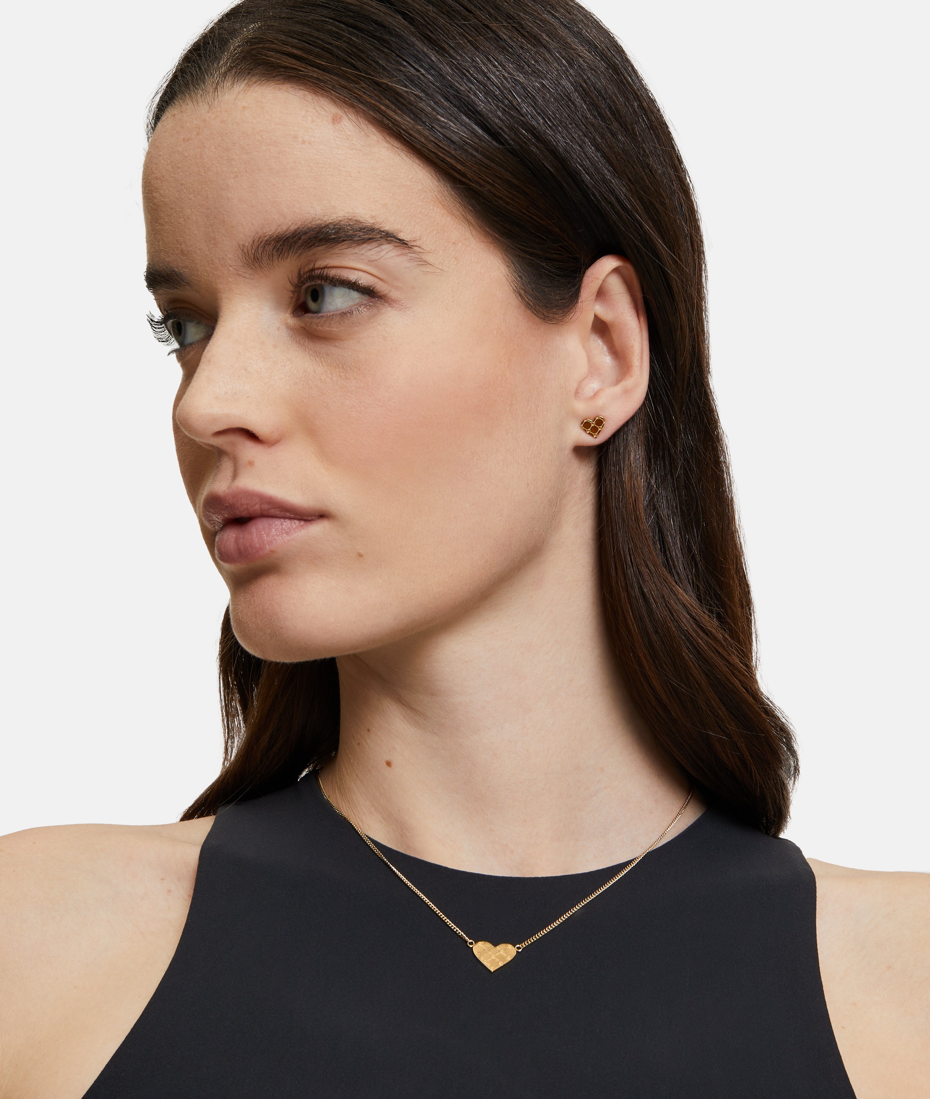 Eine Frau mit langen braunen Haaren trägt ein schwarzes Top, eine goldene Herzkette und den Tamaris Ohrstecker - The T-Pattern Heart von Tamaris - perfekt als Geschenk.