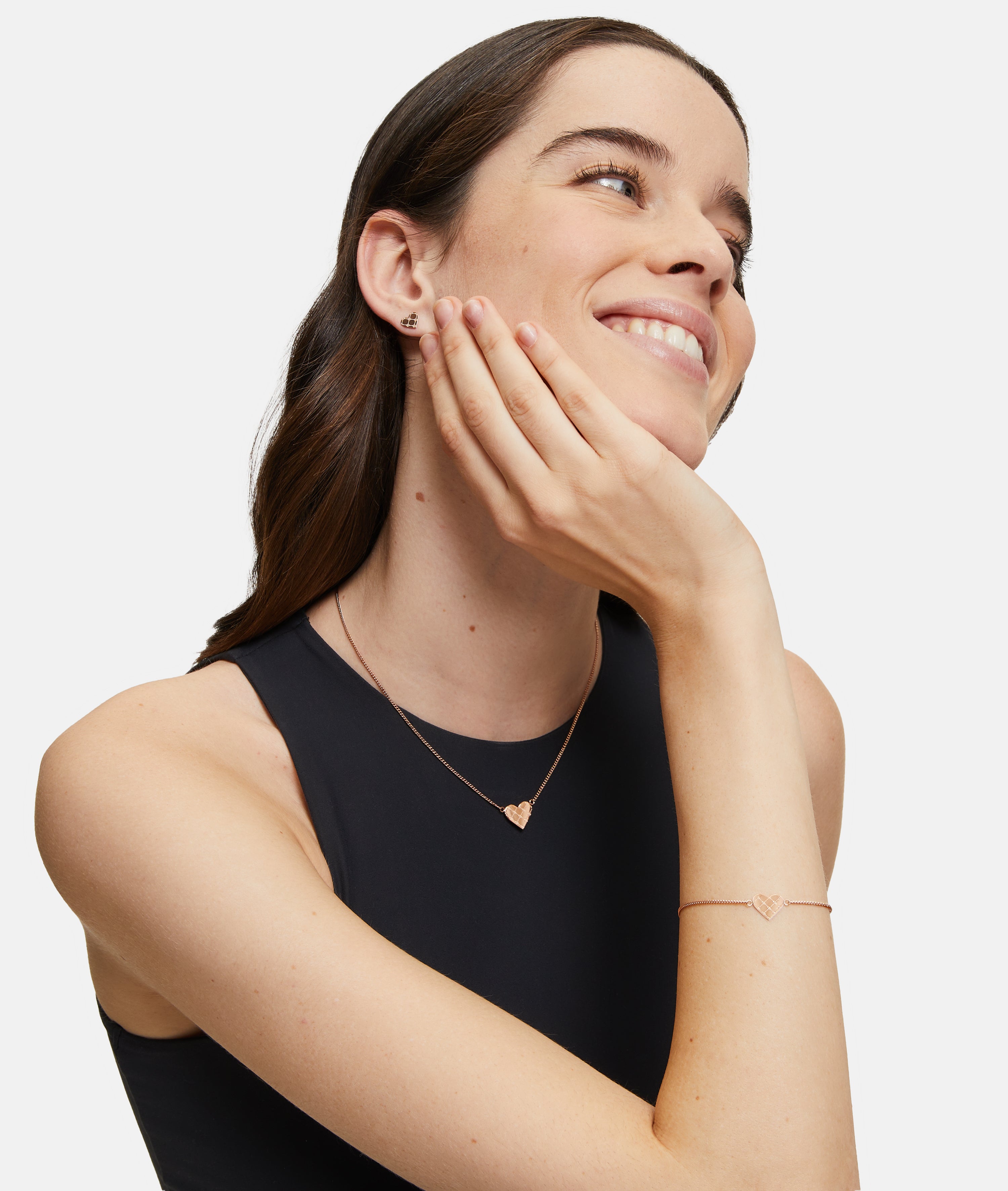 Eine lächelnde Frau trägt die Tamaris Halskette - The T-Pattern Heart mit passendem Armband und Ohrringen, die Hand sanft auf dem Gesicht - ein schönes Geschenk von Tamaris für einen besonderen Menschen.