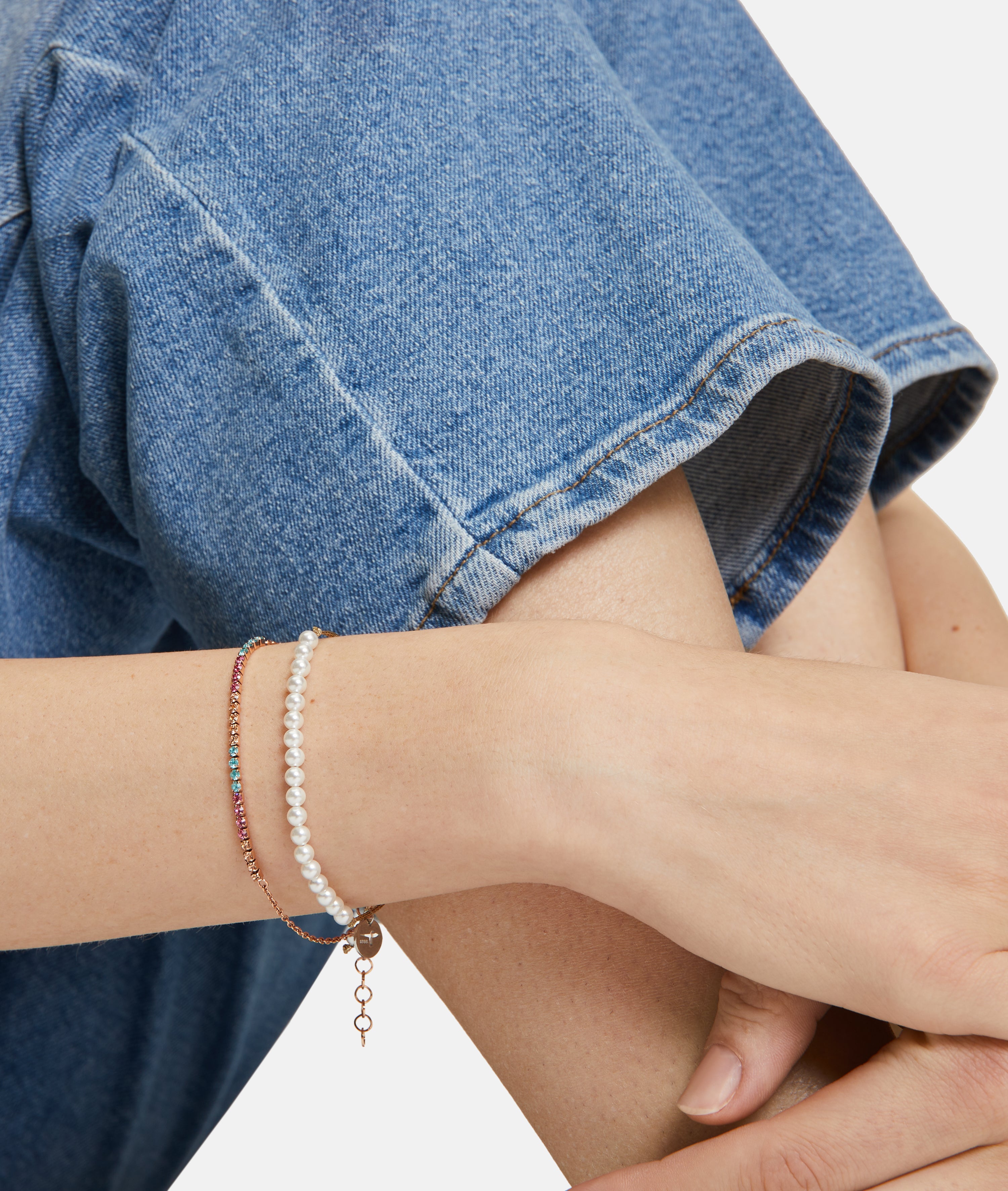 Nahaufnahme einer Person in Blue Jeans, die zwei zarte Armbänder trägt - das eine ist das mit Glasperlen verzierte Armband The Pearl Beads von Tamaris, das andere besteht aus kleinen farbigen Steinen.